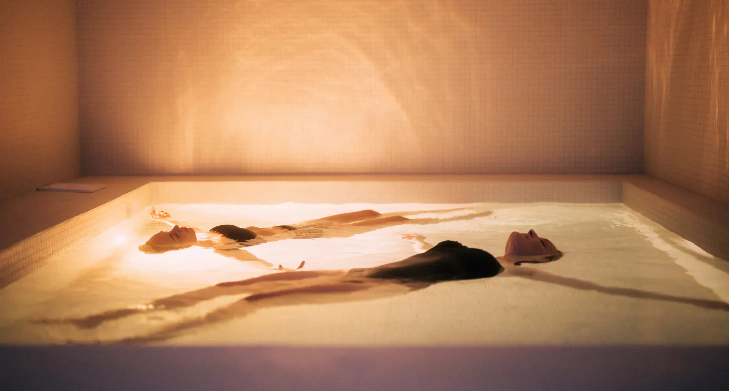 Bassin thermal éclairé de lumière ambrée où deux silhouettes flottent en état de relaxation profonde
