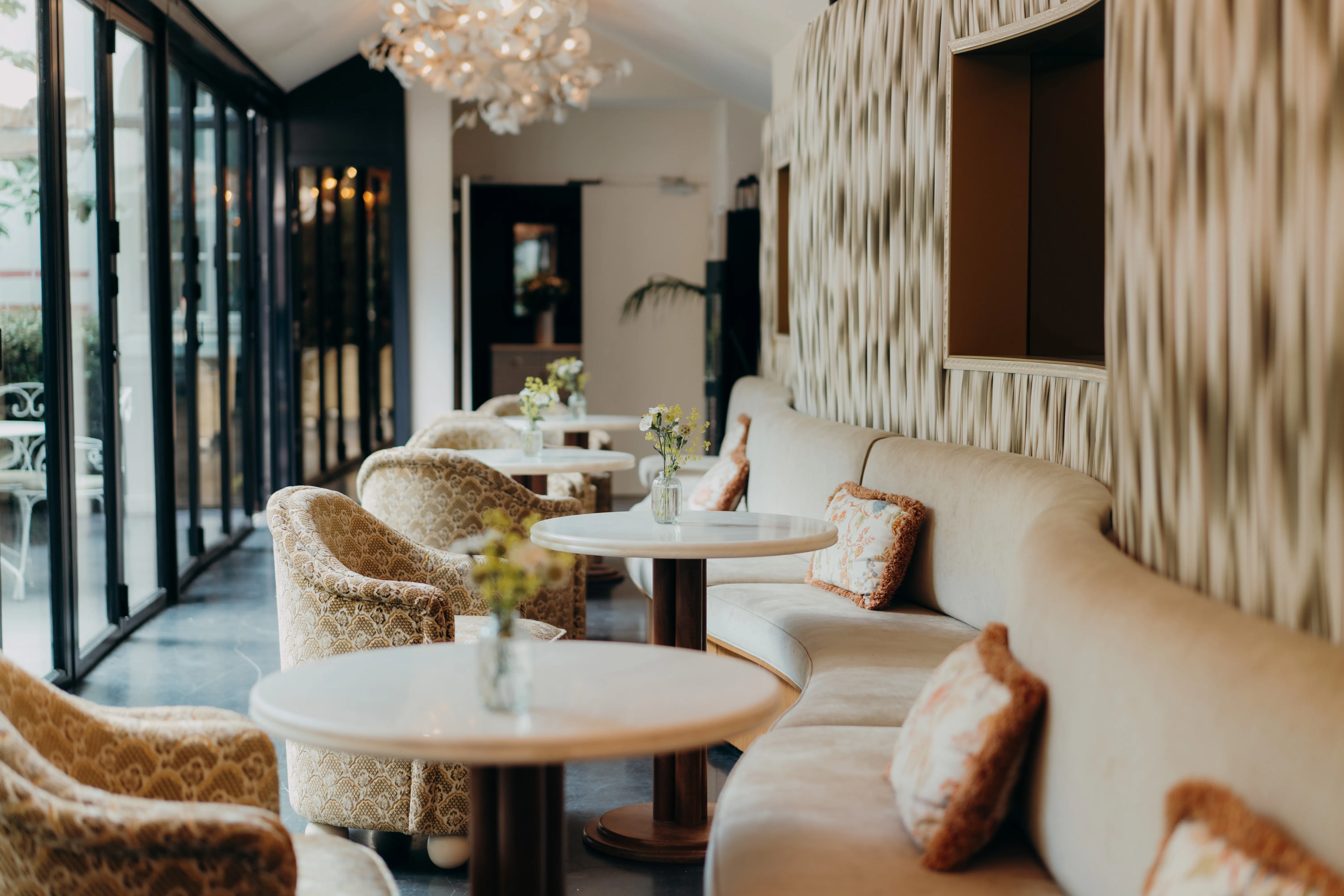 Salon avec banquette beige incurvée, fauteuils à motifs et tables rondes en marbre sous un lustre floral