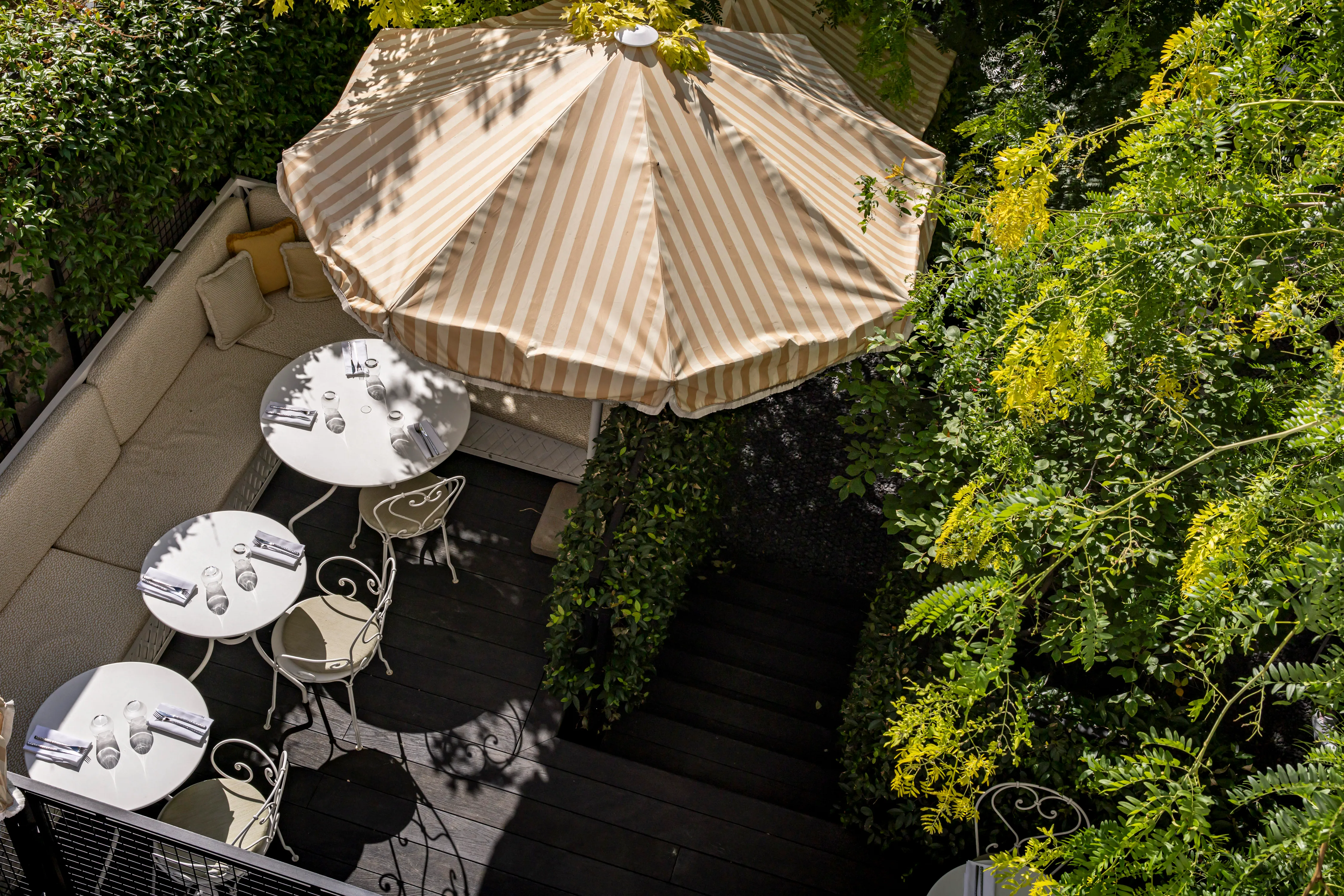 Vue aérienne d'une terrasse ombragée avec parasol rayé beige et tables rondes en fer forgé, entourée de glycine