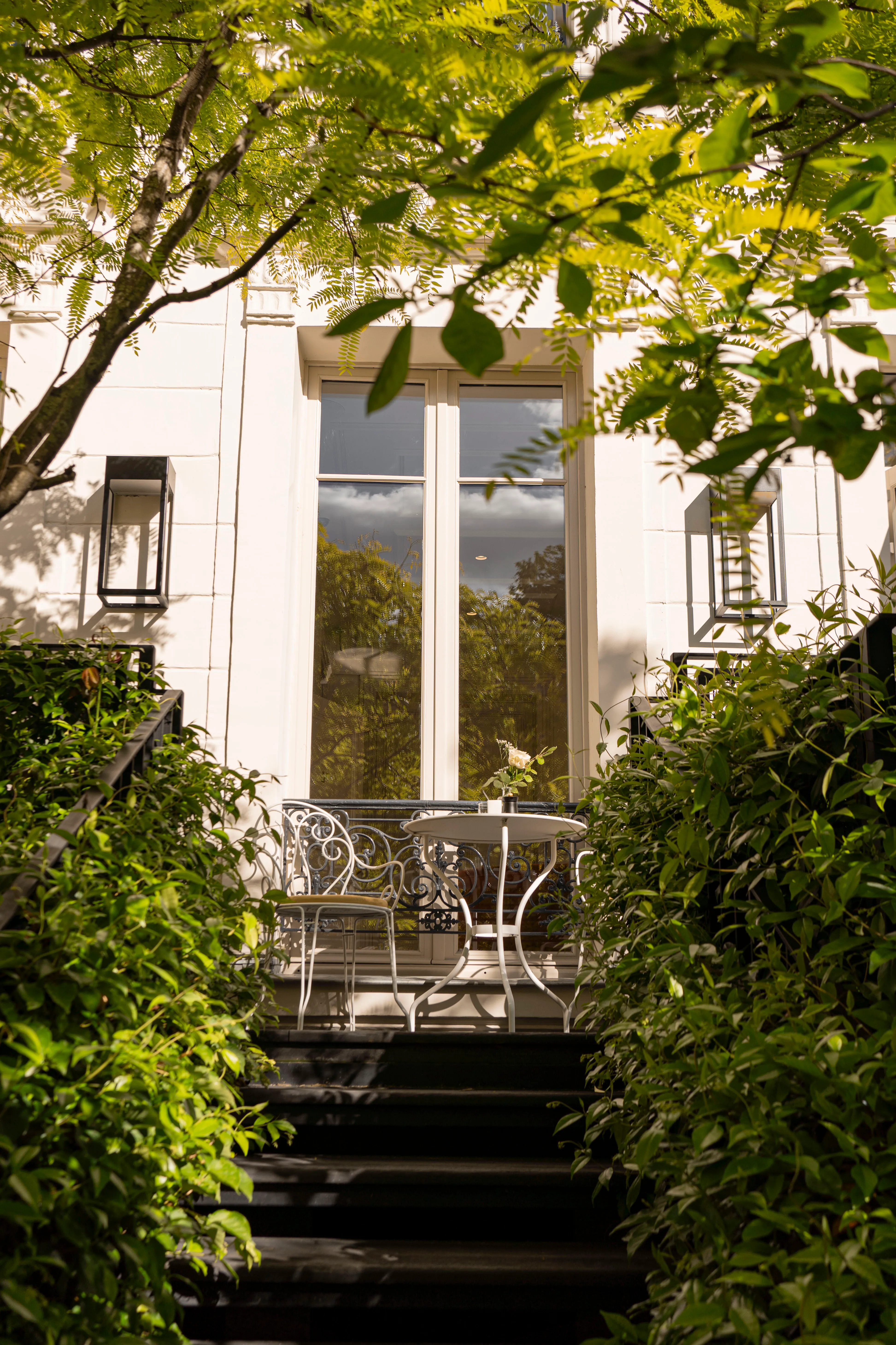 Petit balcon avec table et chaise en fer forgé blanc, entouré de feuillage verdoyant devant une fenêtre