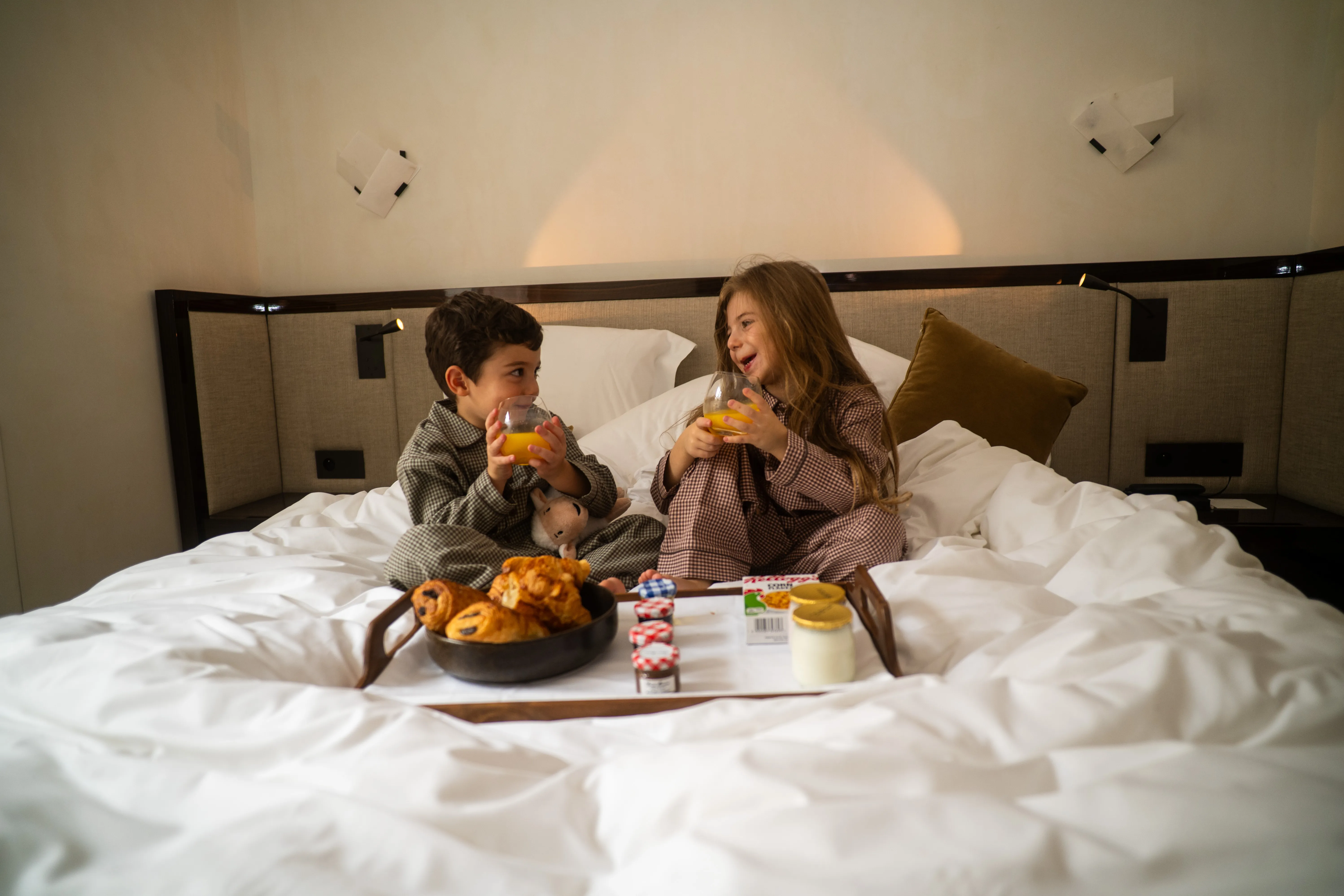 Deux enfants en pyjama riant en buvant du jus d'orange, plateau de viennoiseries et confitures sur le lit