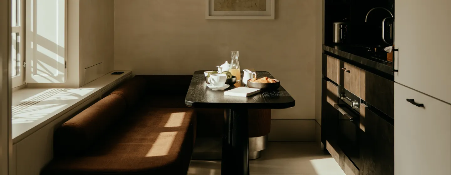 Petit-déjeuner dressé sur une table près d'une fenêtre ensoleillée, avec banquette en velours brun et cuisine en arrière-plan