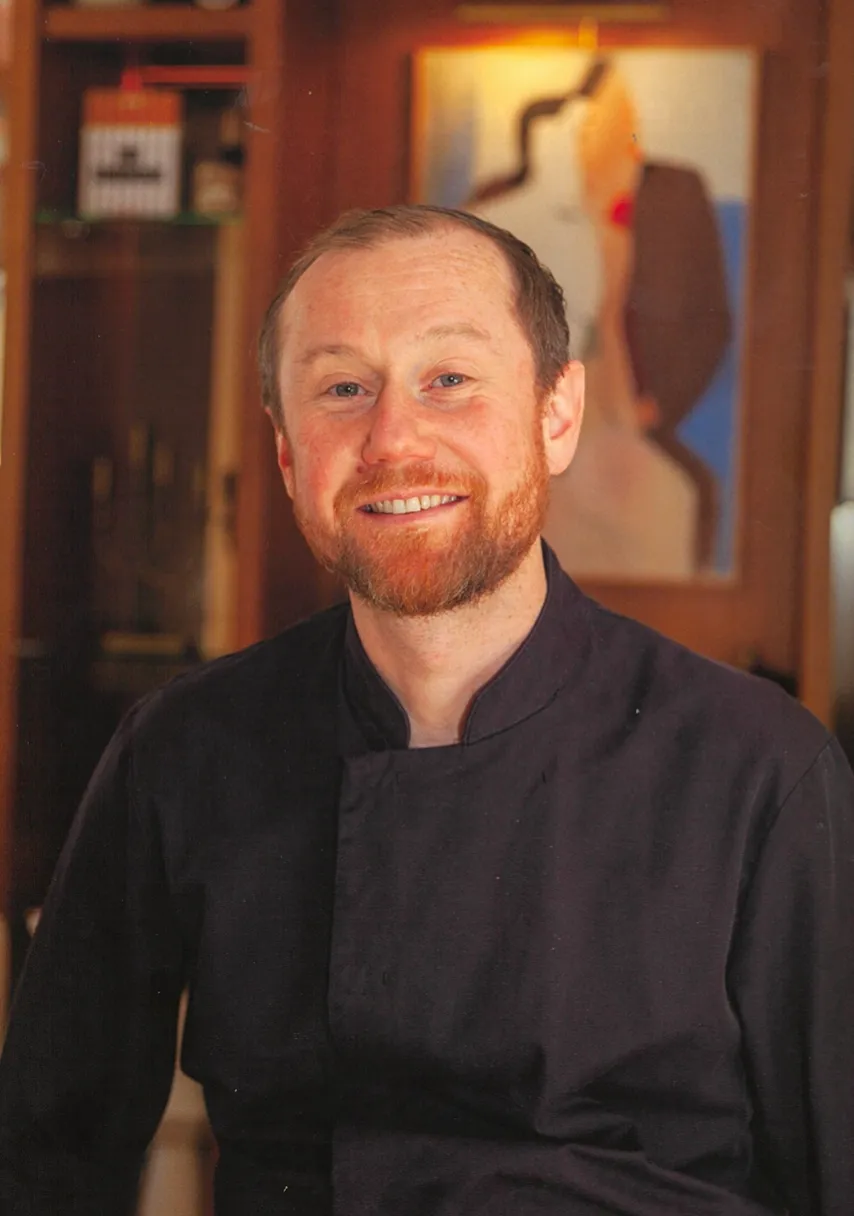 Portrait souriant d'un chef cuisinier roux en veste noire, devant une œuvre d'art murale