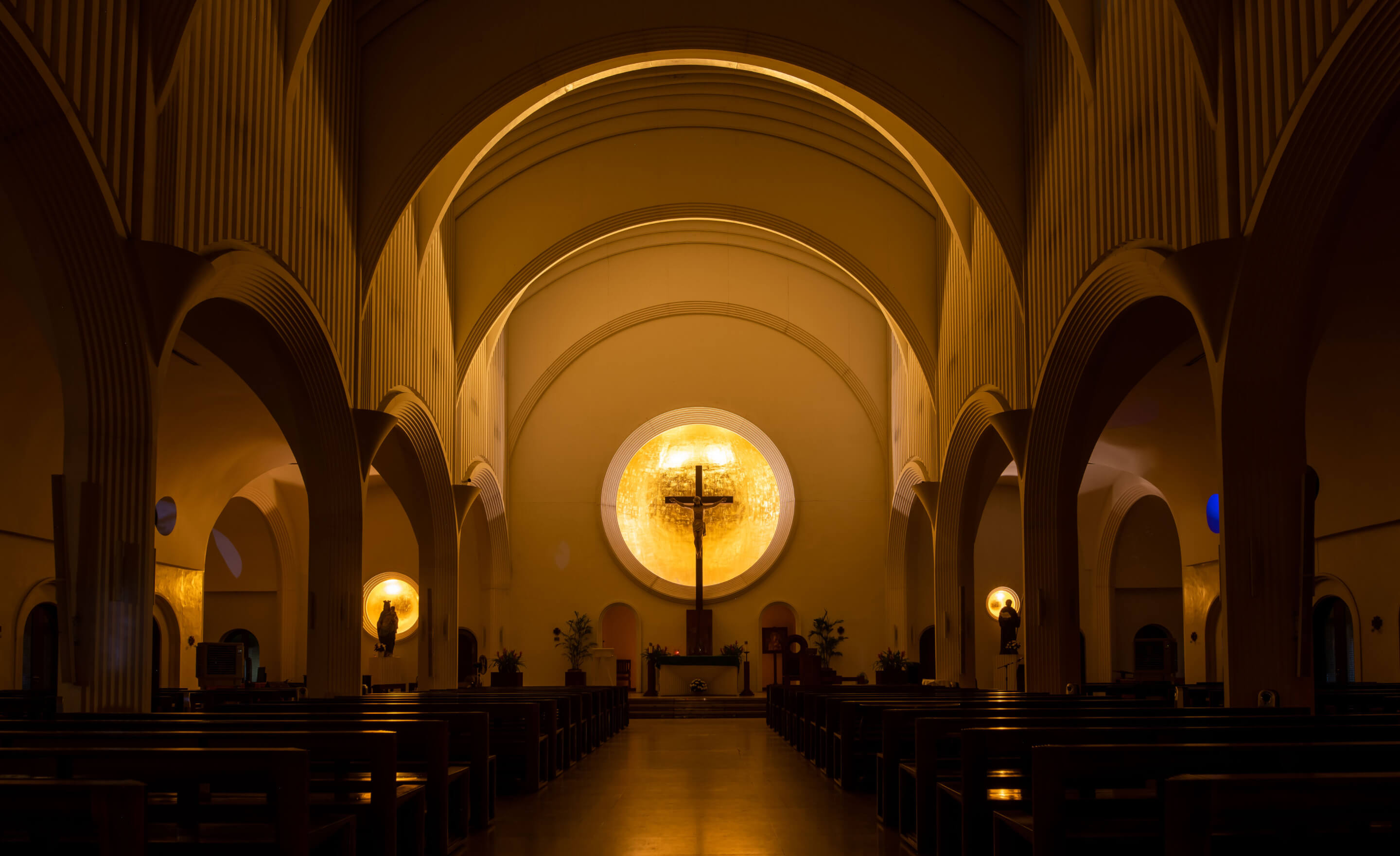 Interior of a church with arched ceilings, wooden pews, and a crucifix illuminated by a circular golden light.