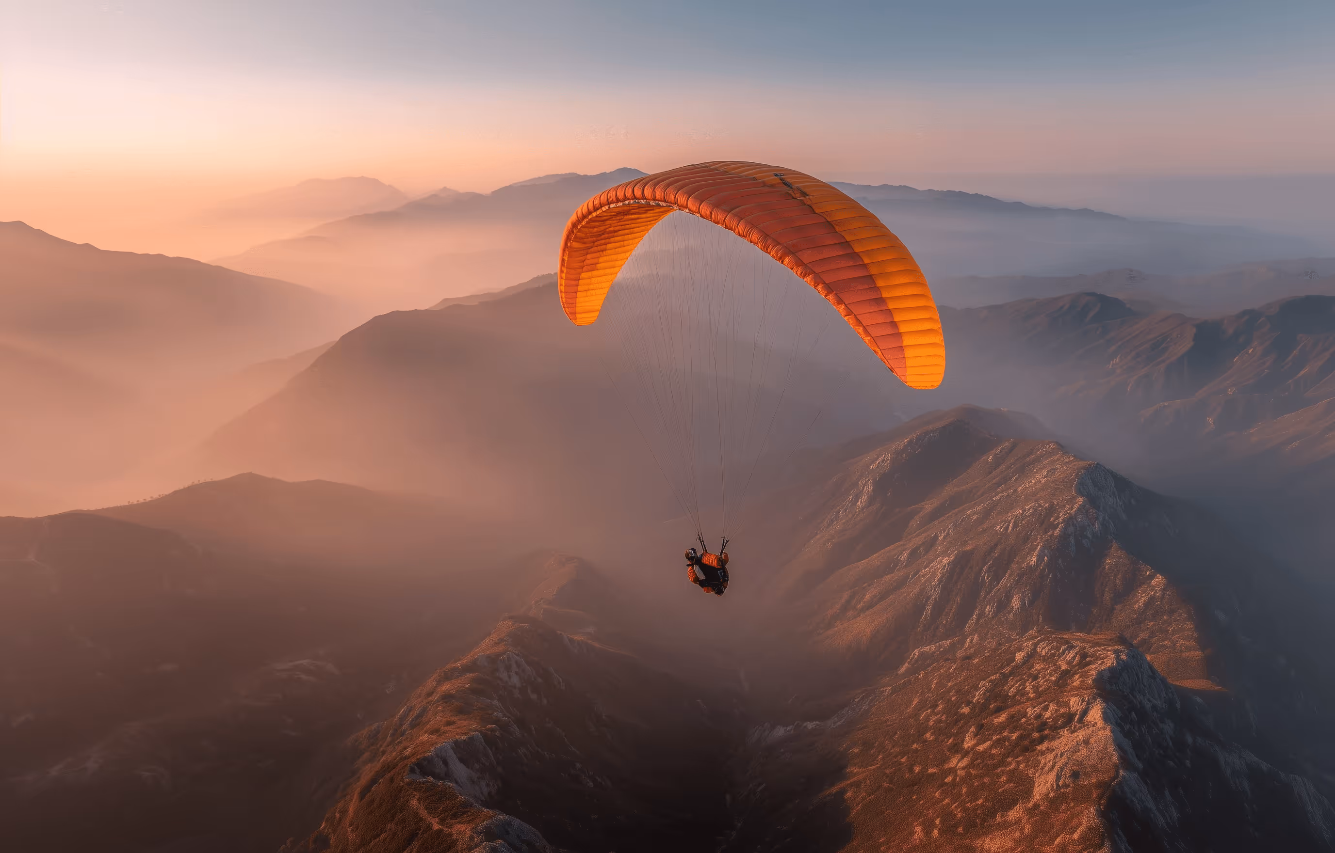 Person paragliding over misty mountains during a warm sunrise or sunset.