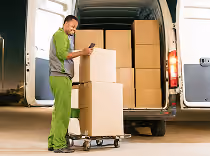 Worker moving stacked cardboard boxes on a hand truck into a white delivery van.