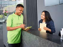 Man in green shirt interacts with woman at a reception desk, both smiling and holding tablets.