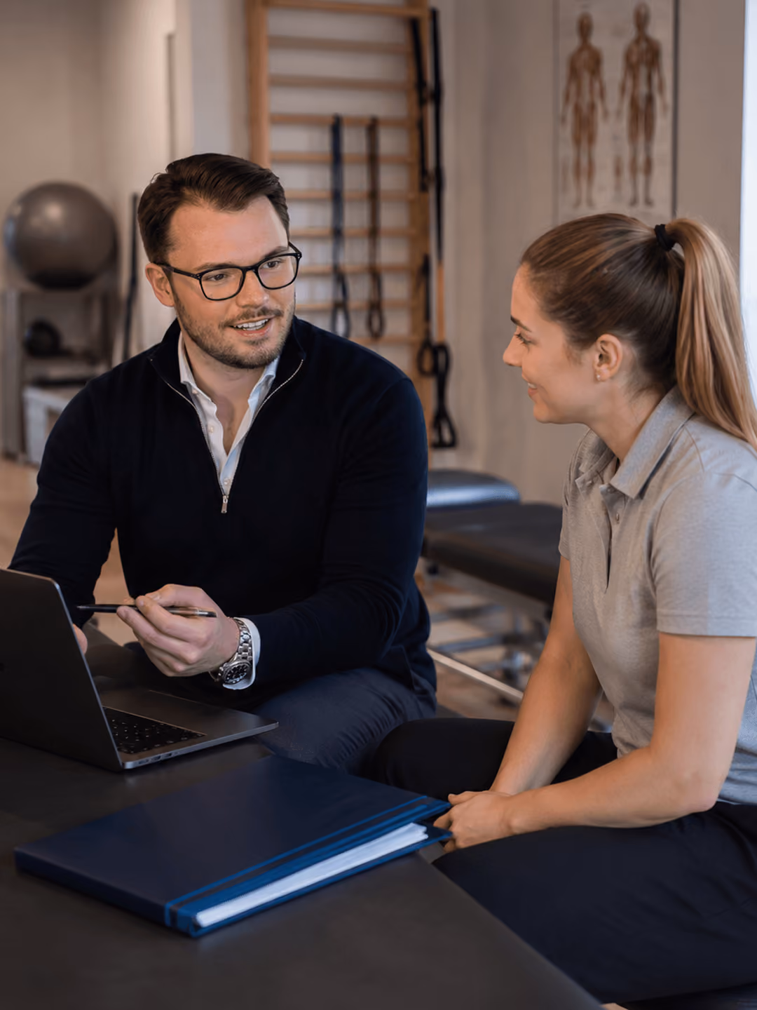 Mann im Gespräch mit Frau in moderner Physiotherapiepraxis, Laptop und Dokumente auf Tisch.