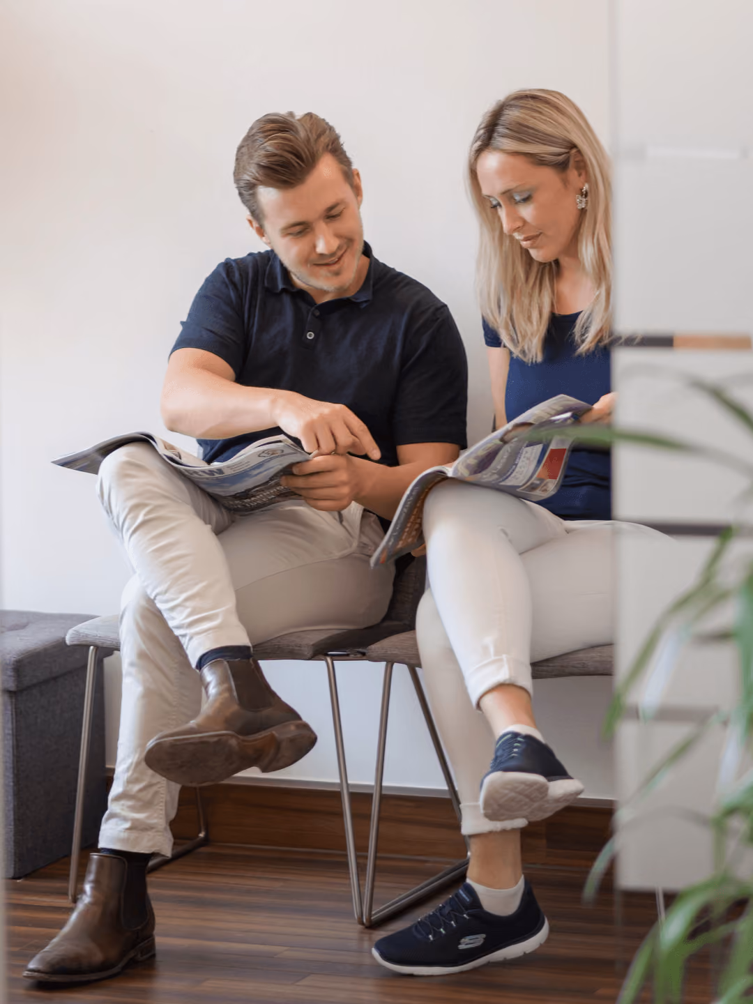 Zwei Personen lesen gemeinsam Zeitschriften auf einer Bank, beide entspannt gekleidet, helle Umgebung.