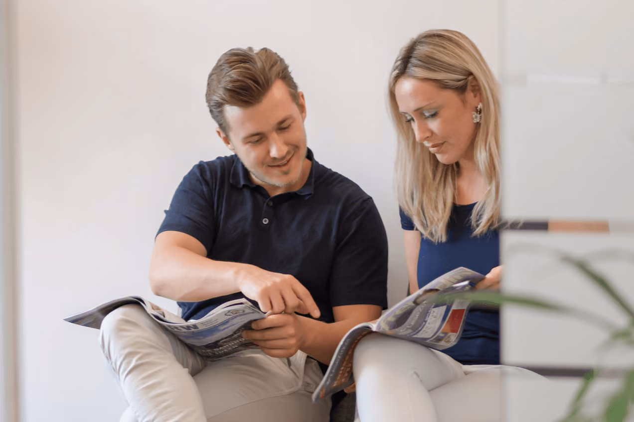 Zwei lächelnde Personen sitzen und lesen Zeitschriften; entspanntes Indoor-Setting.