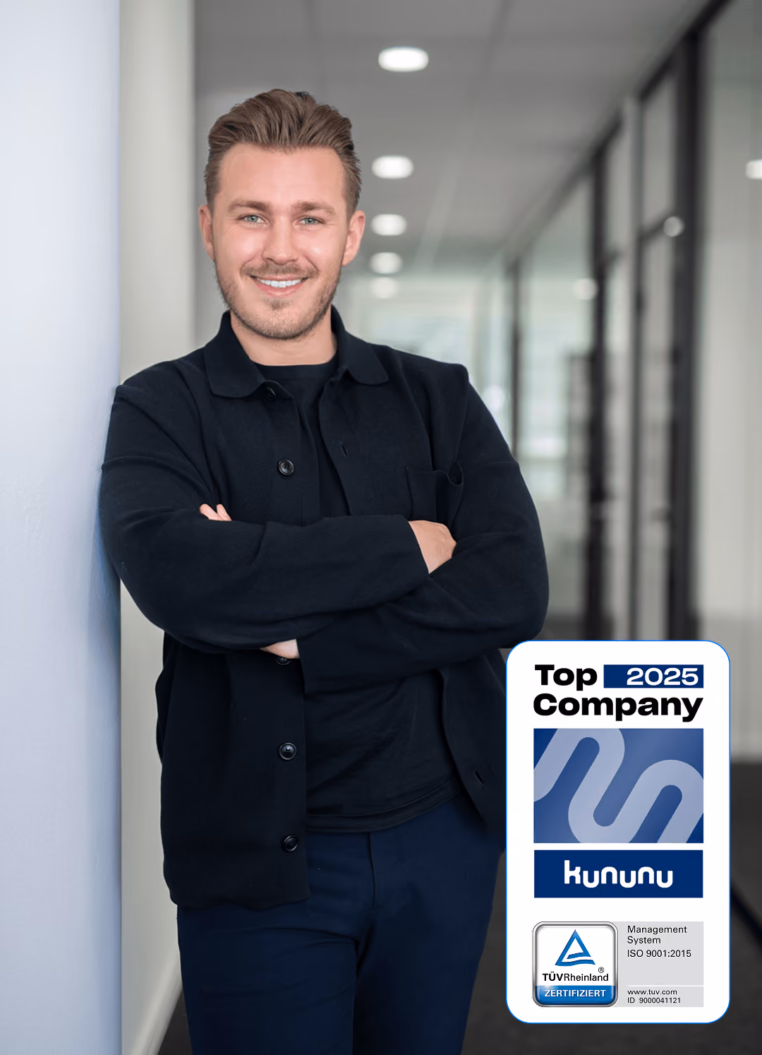 Mann in schwarzer Kleidung lehnt lächelnd an weißer Wand in modernem Büro, hält Kununu-Zertifikat "Top Company 2025".