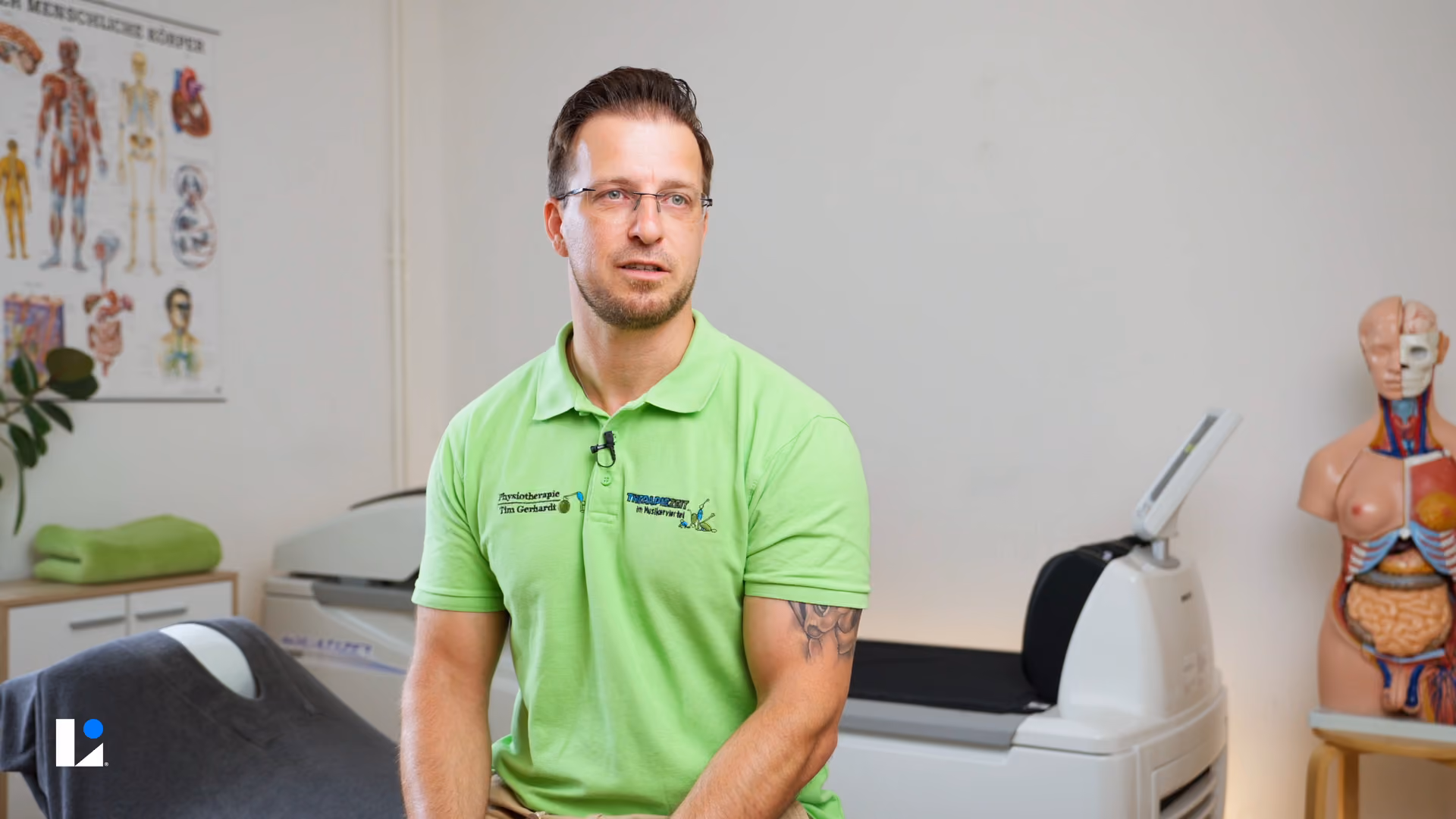 Mann in grünem Polohemd in Physiotherapiepraxis, sitzend neben medizinischen Geräten und Anatomiemodell.
