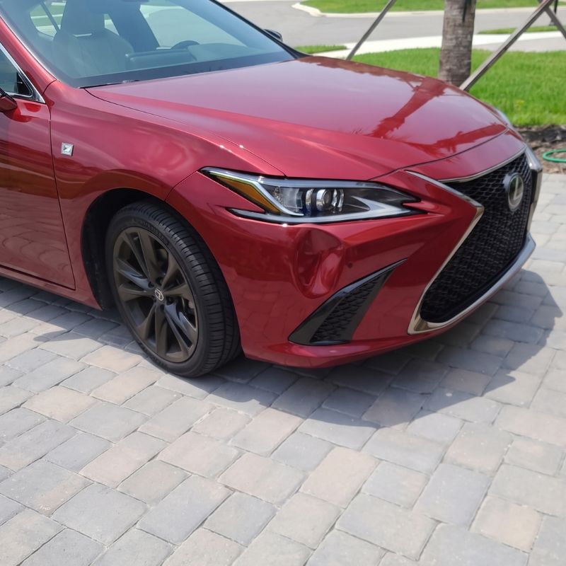 Red Lexus car with a dent on the front right bumper near the headlight parked on a stone driveway.