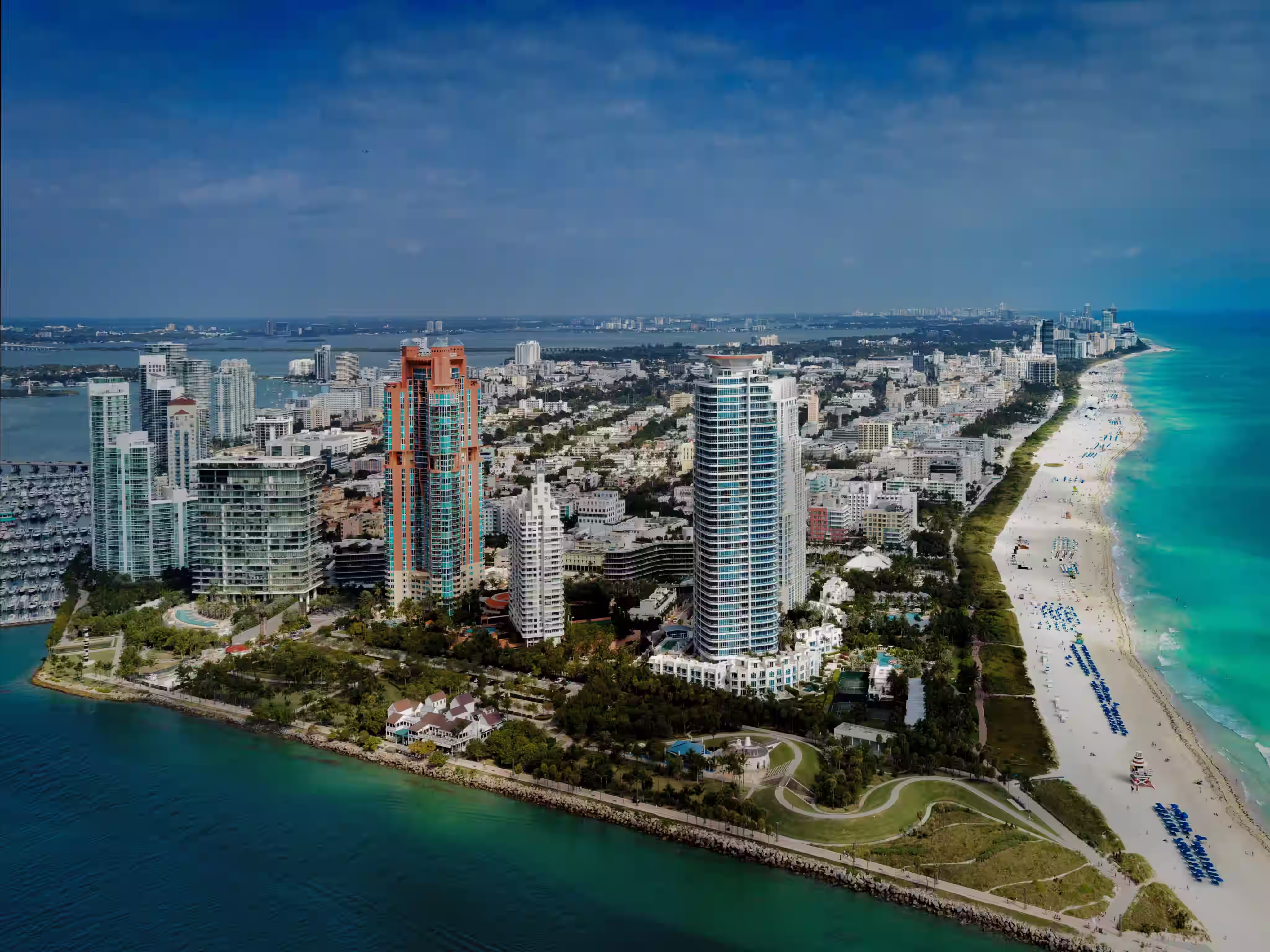 Aerial view of a coastal city with tall buildings, a green park area, and a long sandy beach alongside turquoise ocean water.