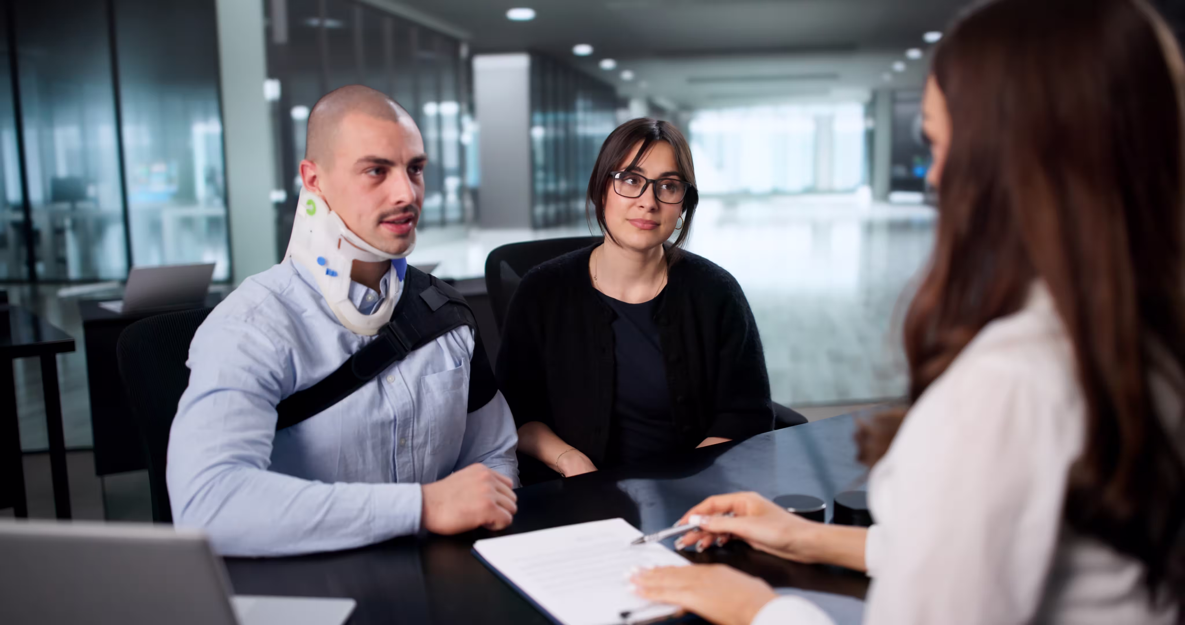 Man with neck brace and shoulder support sitting with woman at a desk, talking to a professional holding a pen over a document.