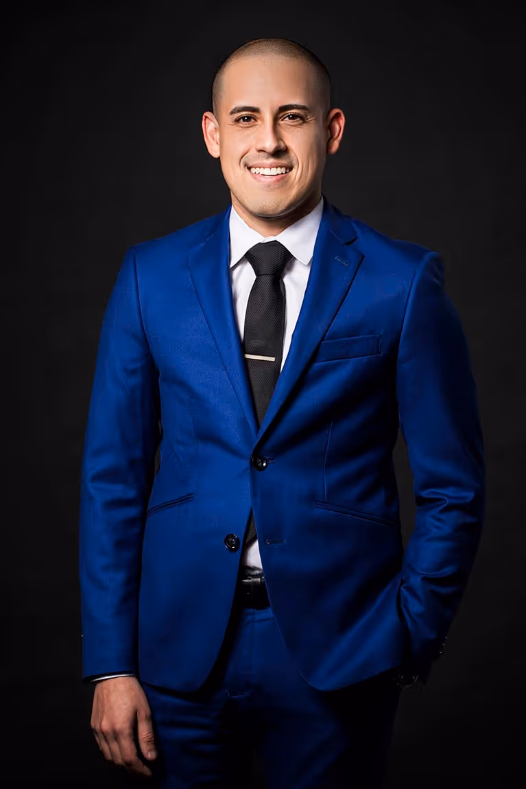 Smiling man wearing a blue suit, white shirt, and black tie standing against a black background.