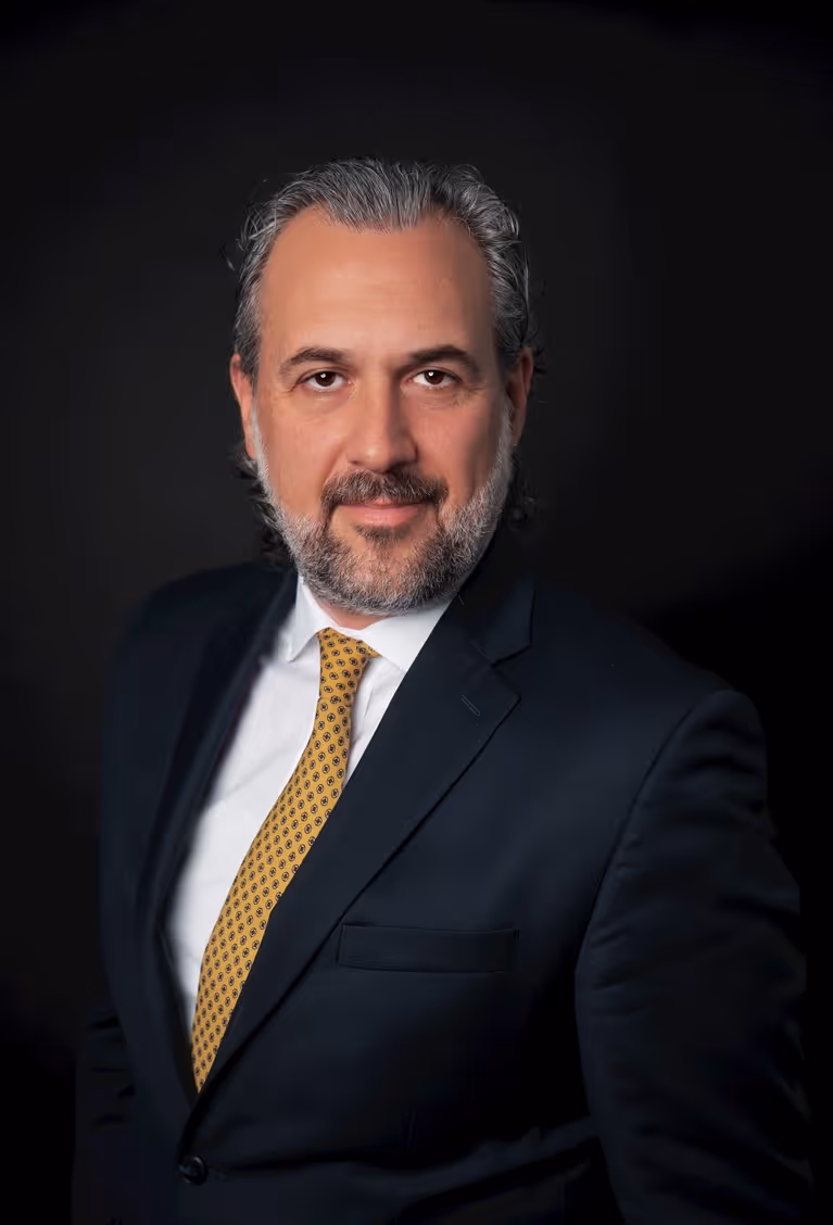 Middle-aged man with gray hair and beard wearing a navy suit, white shirt, and yellow patterned tie against a dark background.