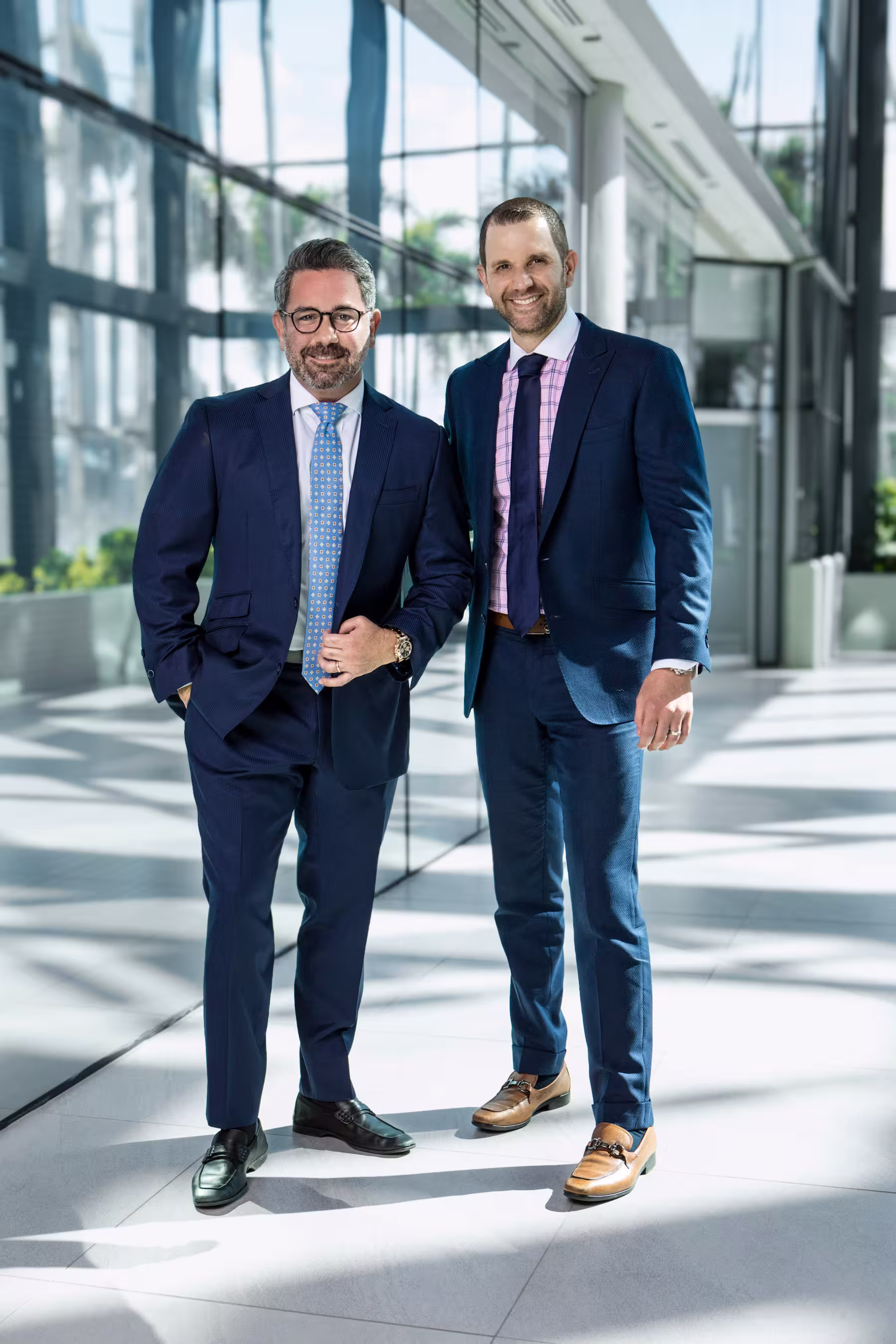 Two businessmen in blue suits standing and smiling inside a modern glass building with natural light.