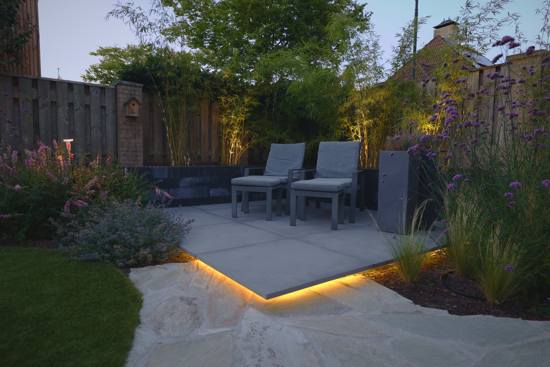 Terras met loungestoelen, sfeerverlichting onder de tegels en bloeiende borders.