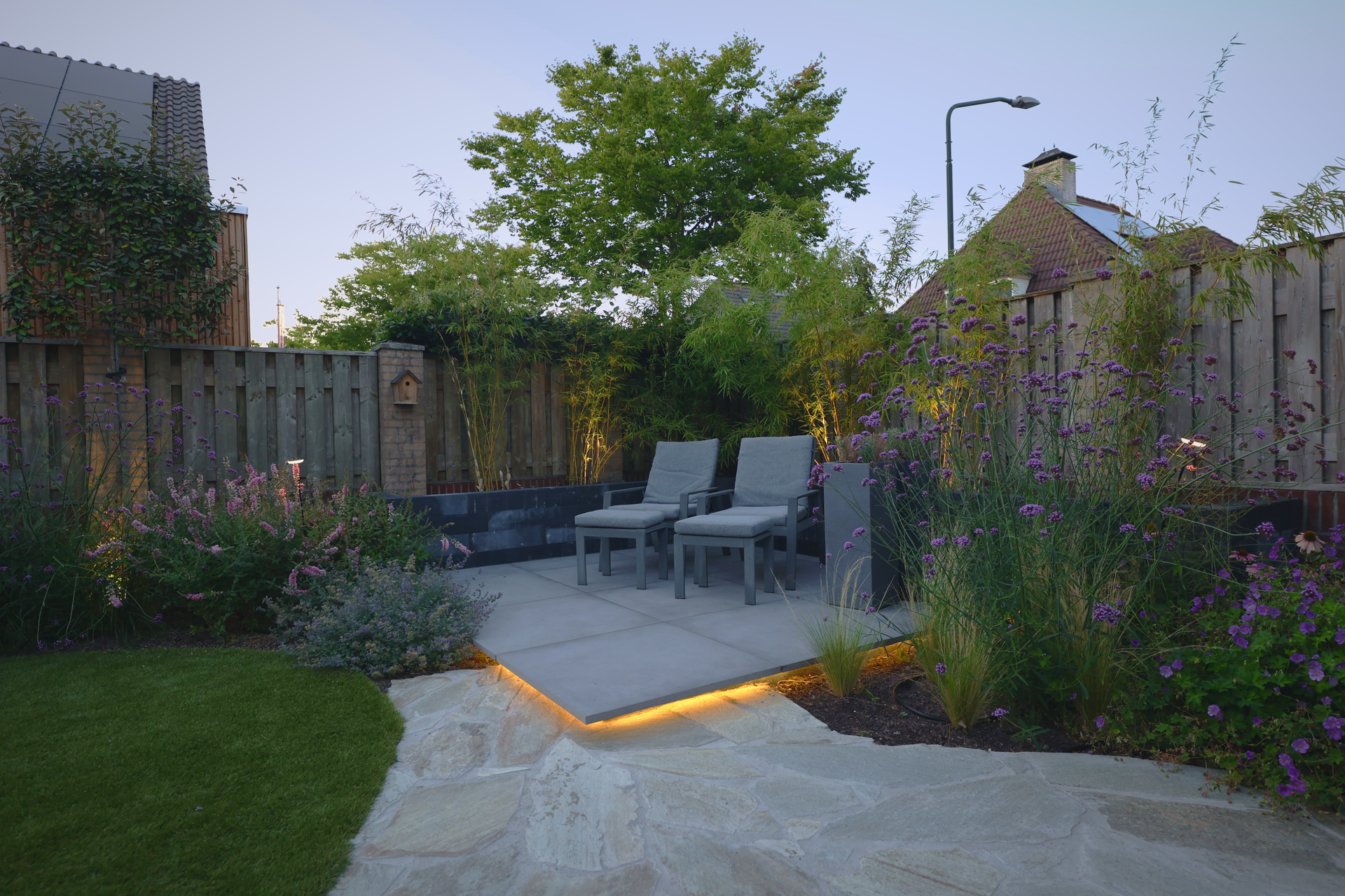 Terras met ligstoelen, natuursteen en verlichte borders in de schemering.