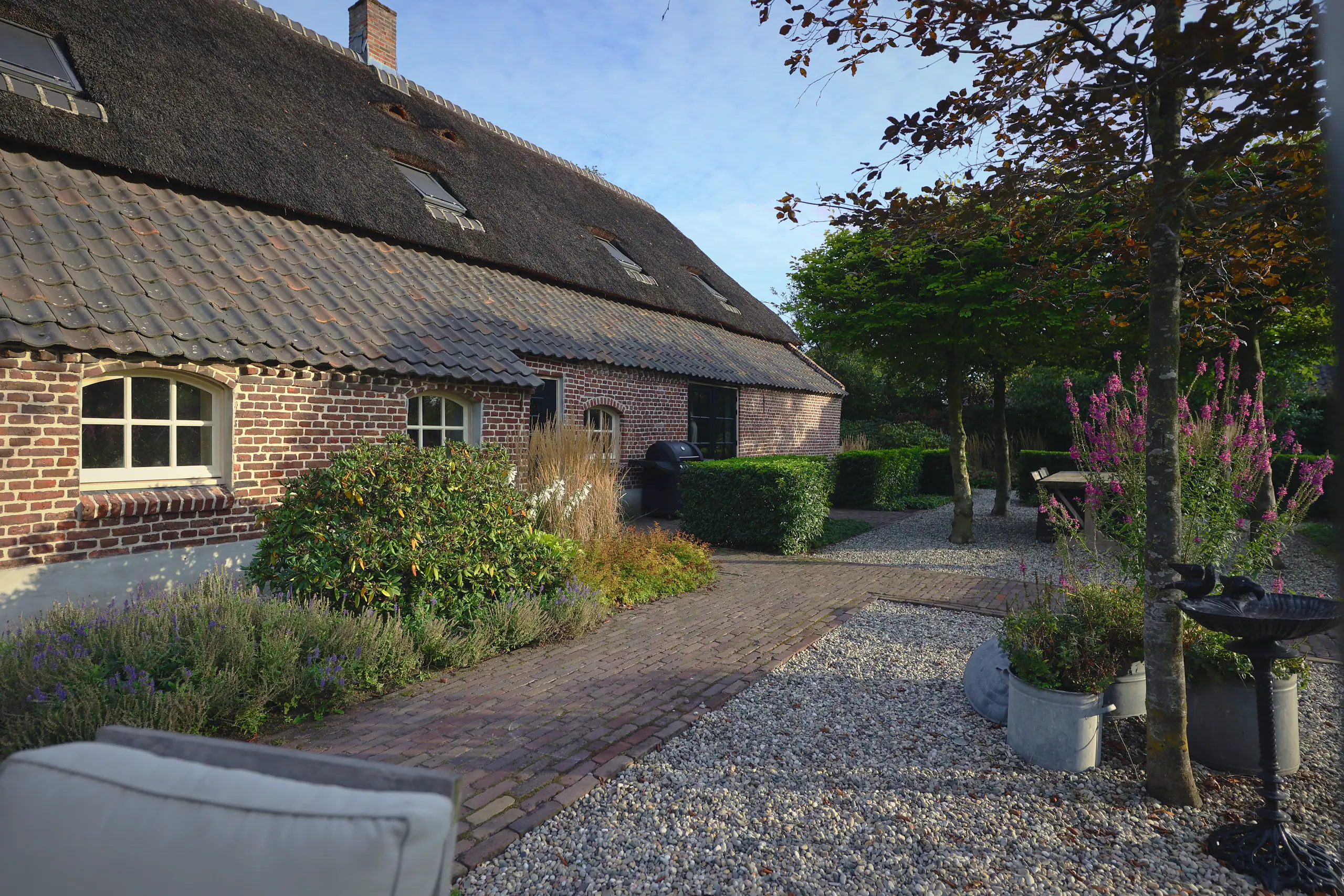 Erf van een rietgedekte boerderij met klinkerpad, grind en beplanting in potten.