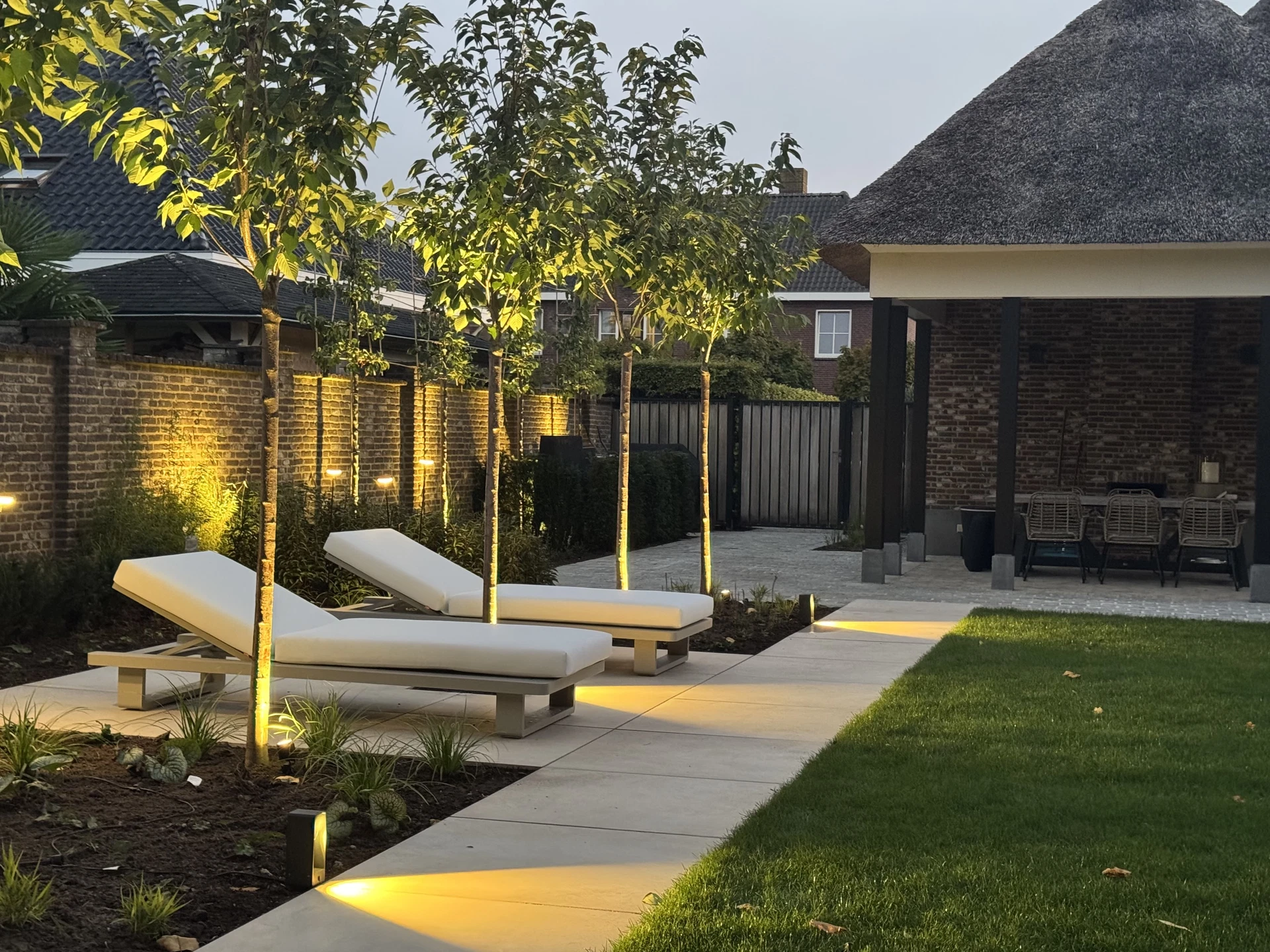 Aangelegde tuin met ligbedden, jonge bomen en grondverlichting langs het terras.