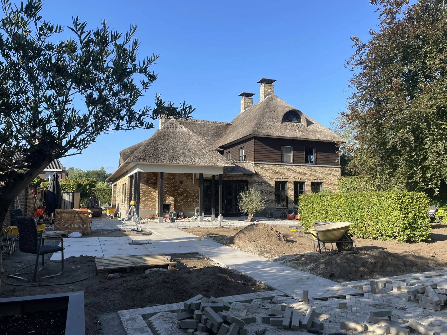 Tuinaanleg in uitvoering bij een rietgedekte villa, met zand en stapels bestrating.