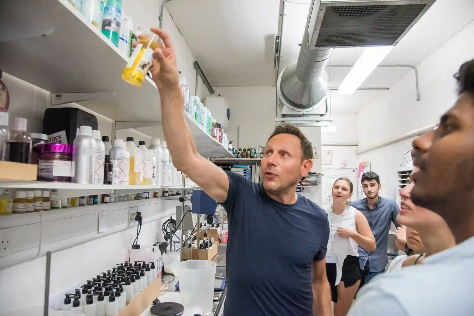 Man in a lab coat holding up a yellow liquid bottle explaining to a group of people in a laboratory.
