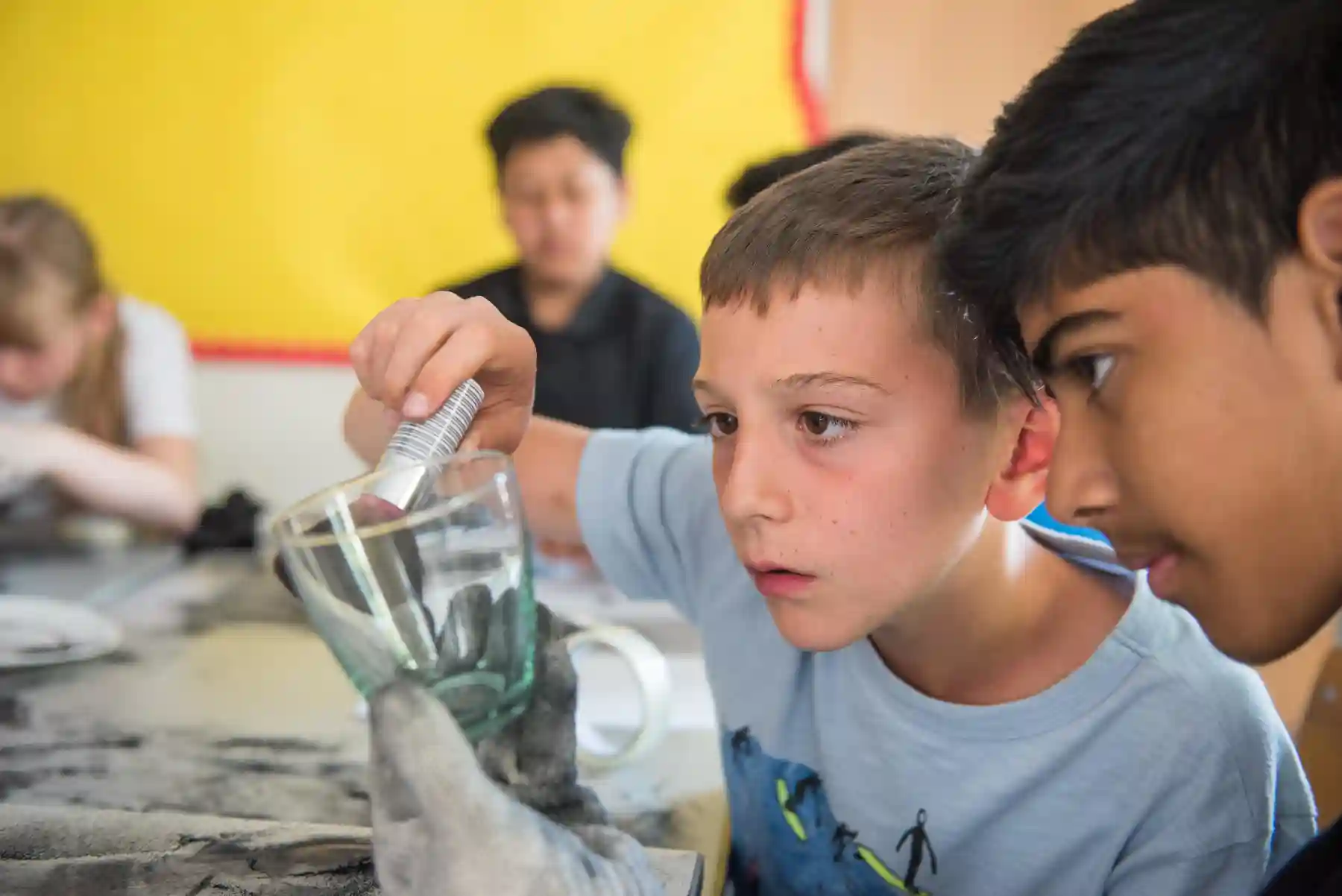 Two boys closely examining a glass measuring cup and a metal tool during a hands-on classroom activity.