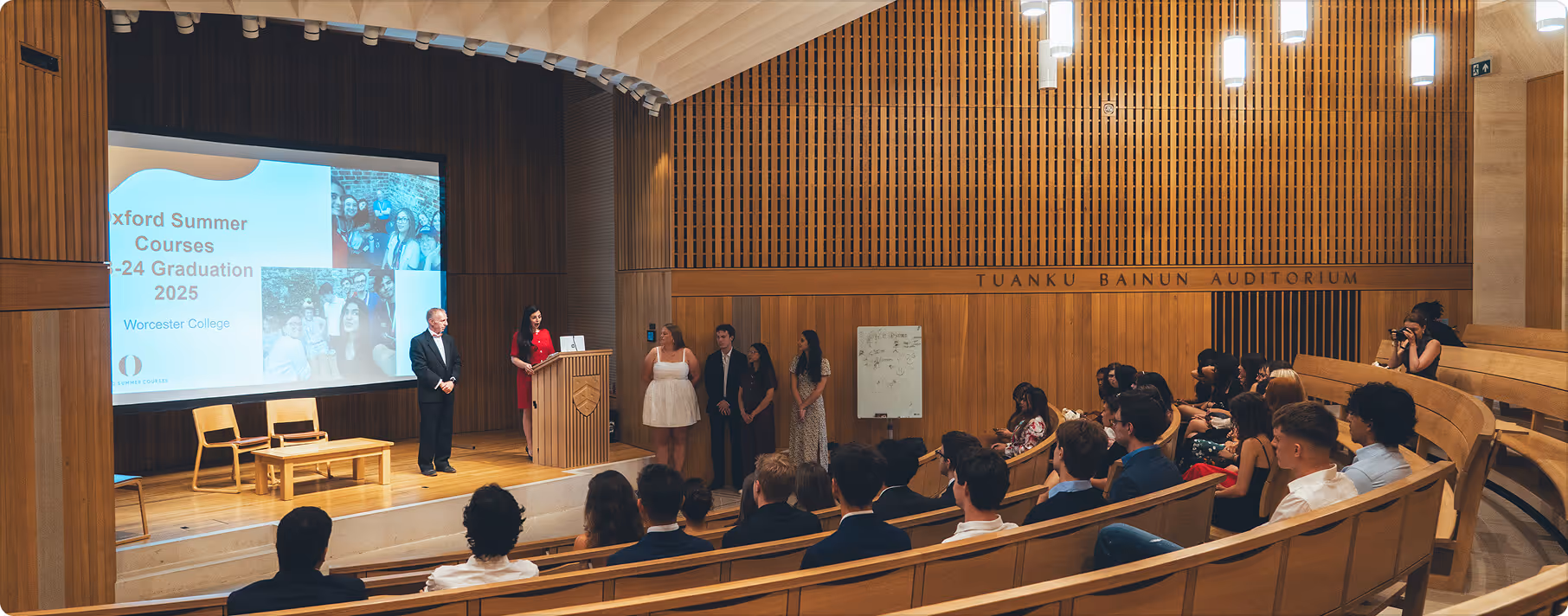 Audience seated in a wooden auditorium watching a graduation ceremony with speakers at a podium and a presentation screen.