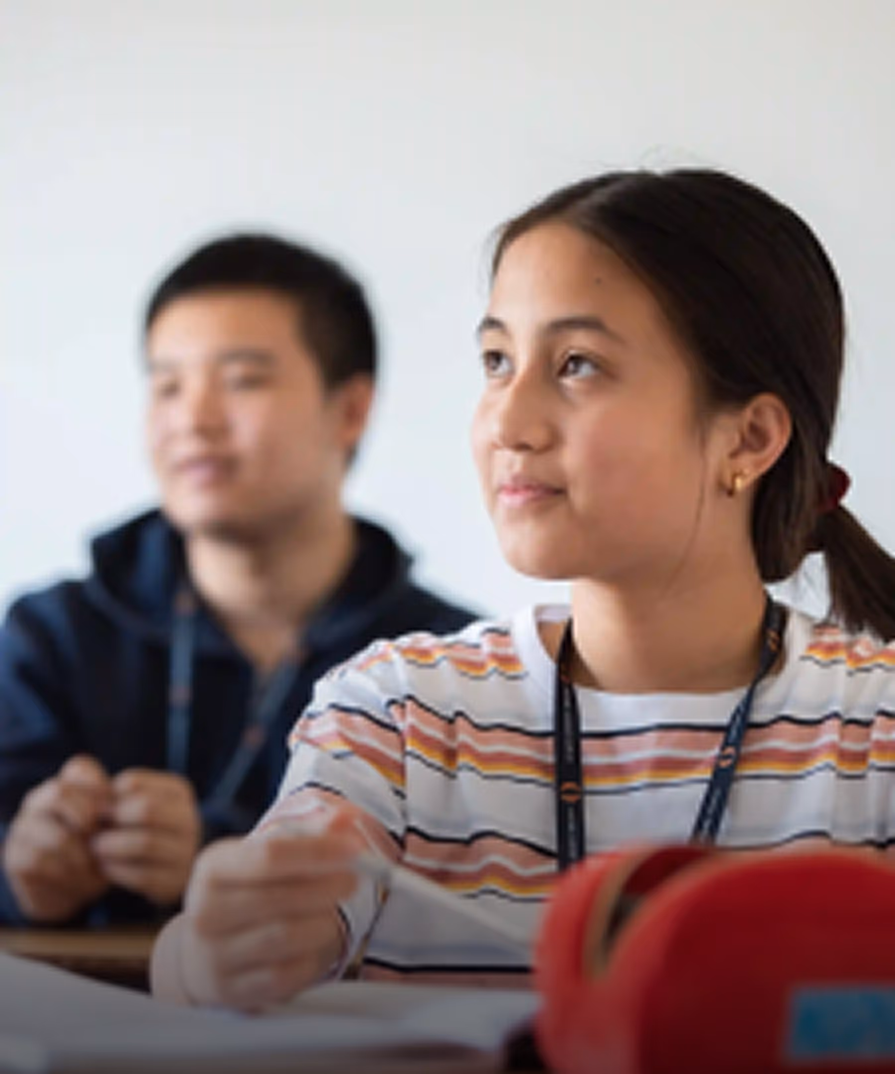 Young female student in a striped shirt attentively looking ahead in a classroom, with a male student blurred in the background.