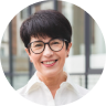 Smiling middle-aged Asian woman with short black hair and glasses wearing a white shirt in a bright indoor setting.