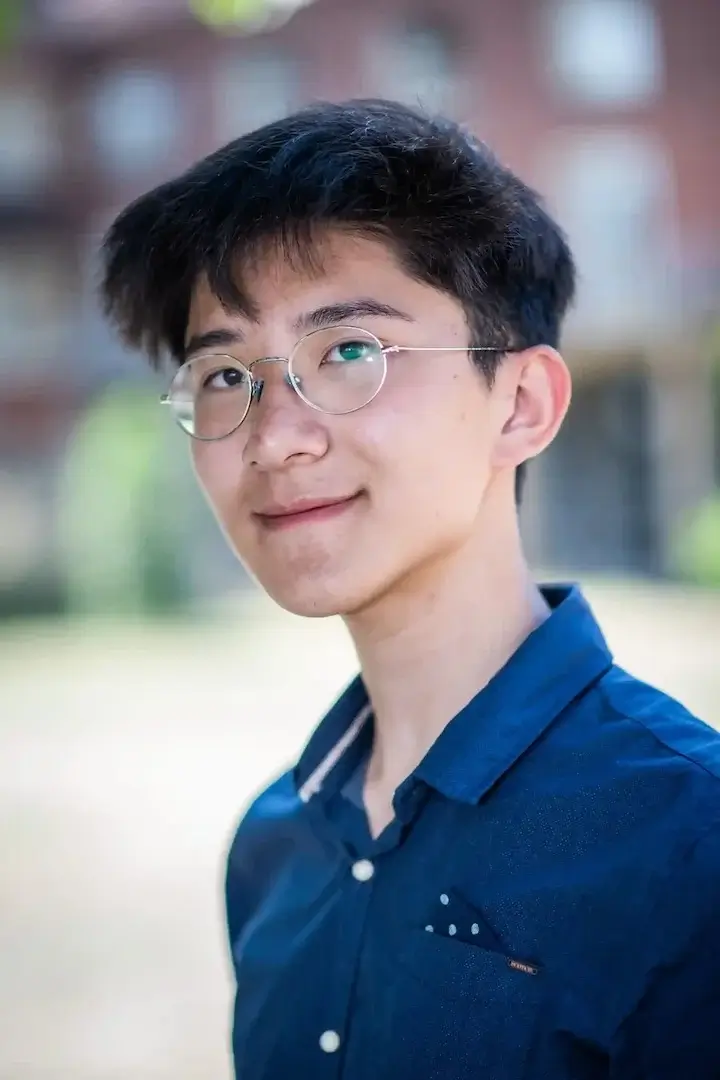 Young man with glasses wearing a blue shirt smiling outdoors with a blurred background.