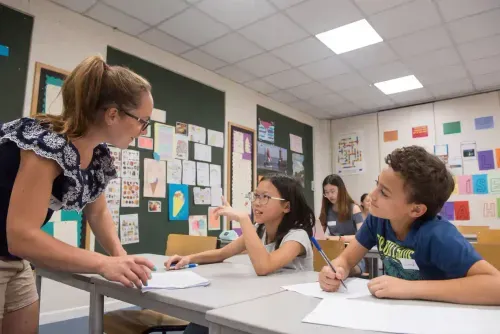 Teacher leaning over a desk talking to two students who are writing in a classroom.