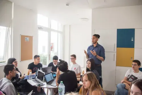 Young man standing and speaking to a group of diverse people seated around a table with laptops in a bright meeting room.