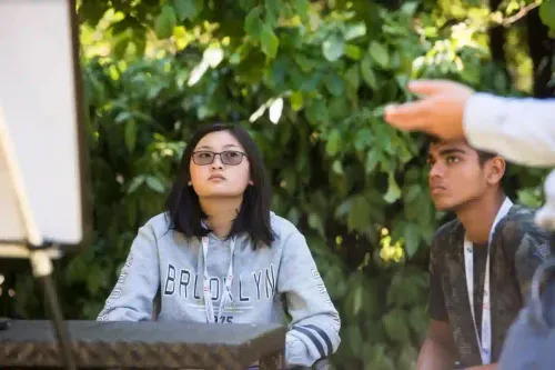 Two young people, a girl wearing glasses and a boy, attentively looking at a person gesturing outside the frame, outdoors with green foliage background.