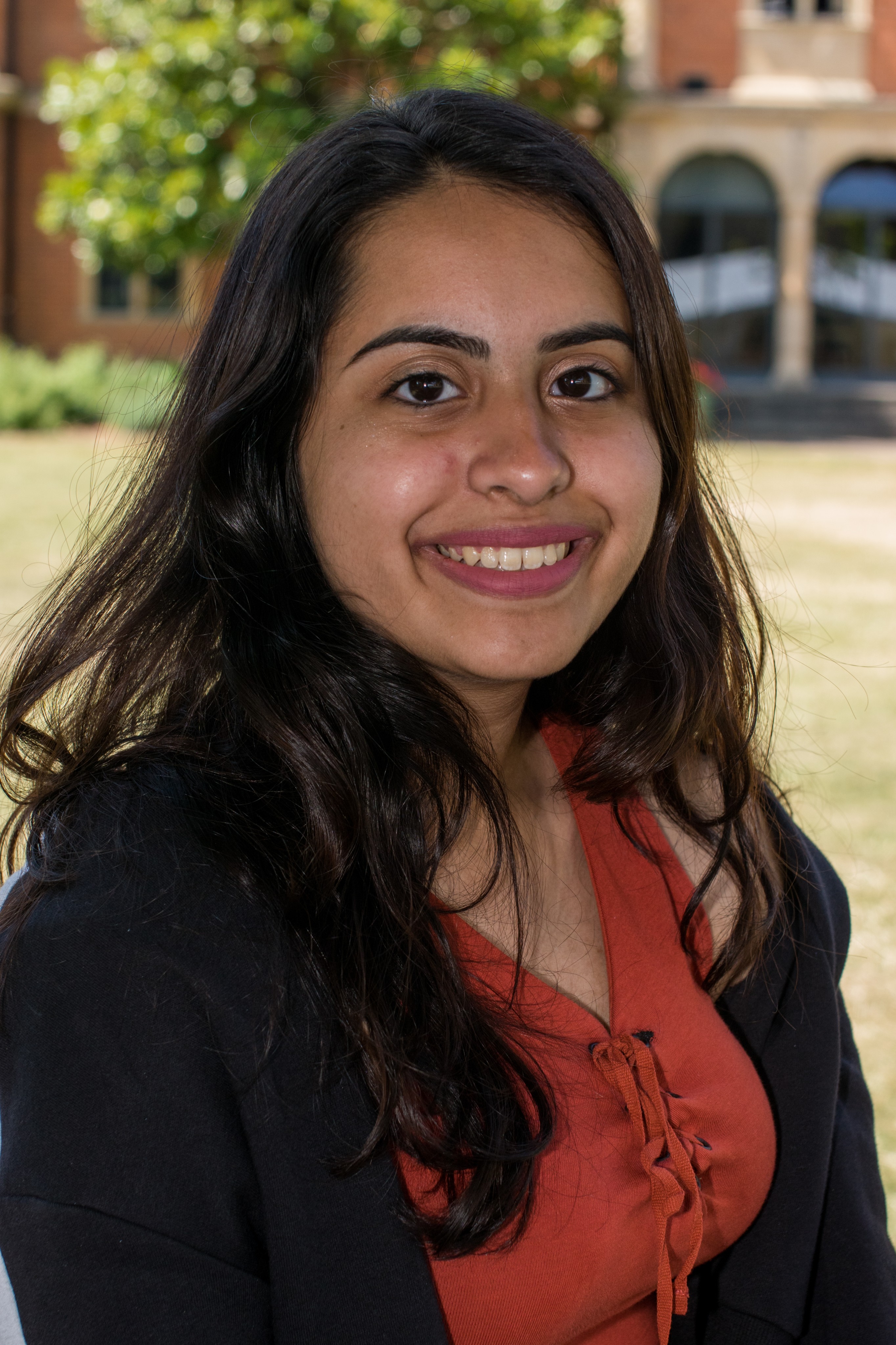 Young woman with long dark hair smiling, wearing a white shirt with black stripes, standing outdoors with blurred green foliage background.