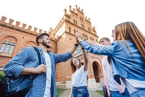 Group of young adults giving a high-five outside a historic brick building with arched windows.