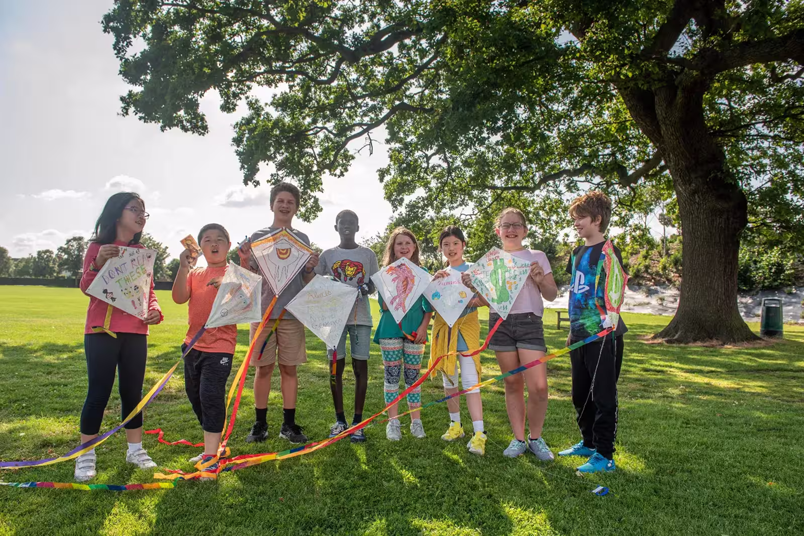 Group of eight diverse children standing on grass under a large tree, holding colorful hand-decorated kites.