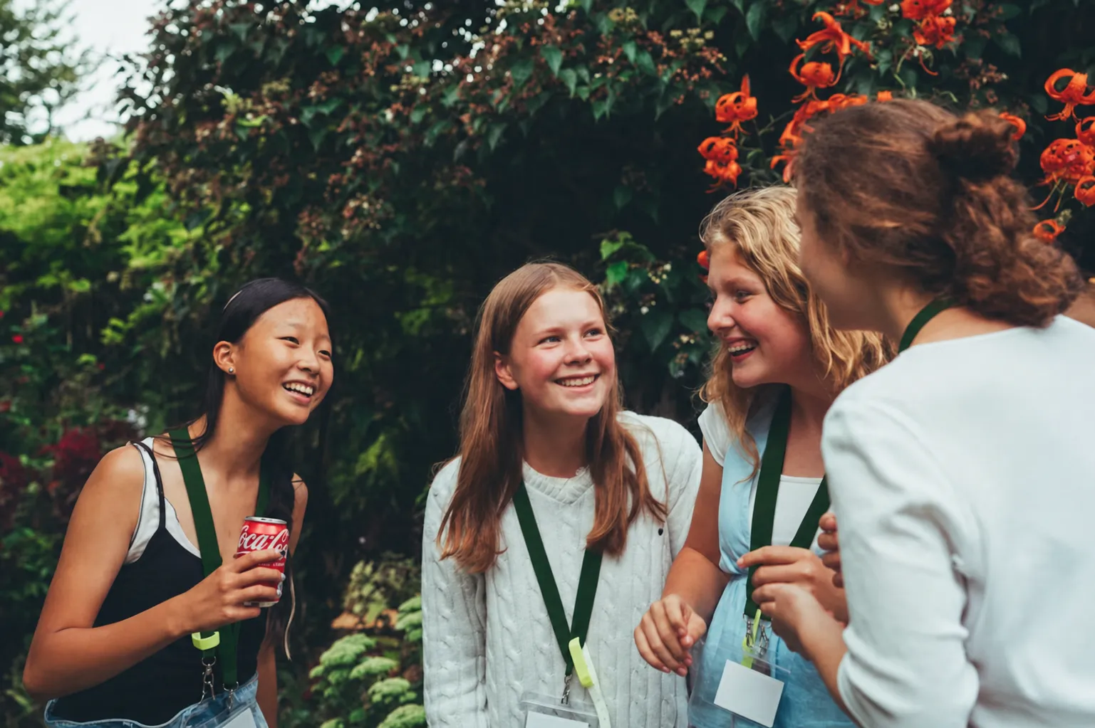 Four teenage girls smiling and chatting outdoors with greenery and orange flowers in the background.