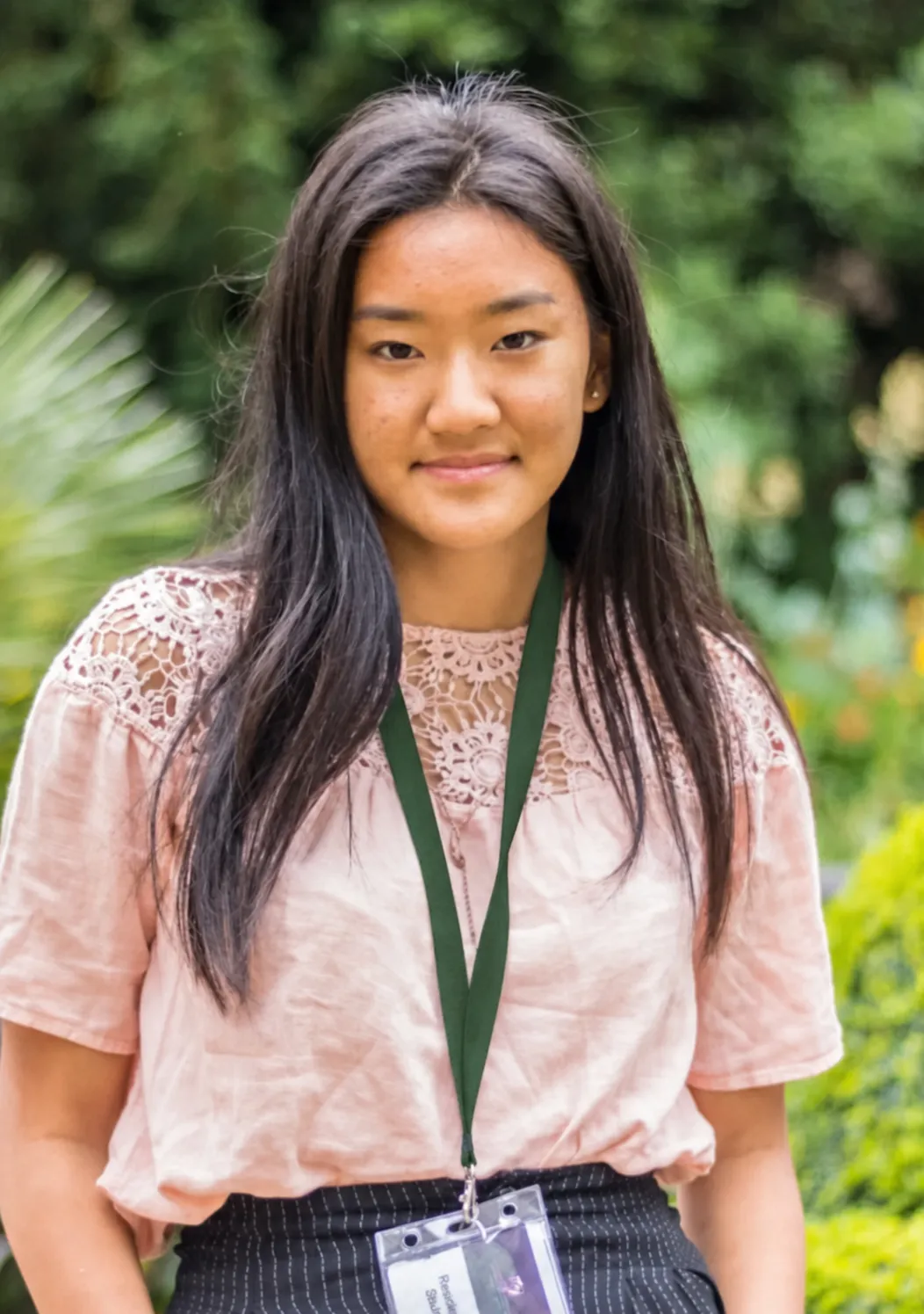 Young woman with long dark hair wearing a pink lace top and a lanyard with an ID badge, standing outdoors.