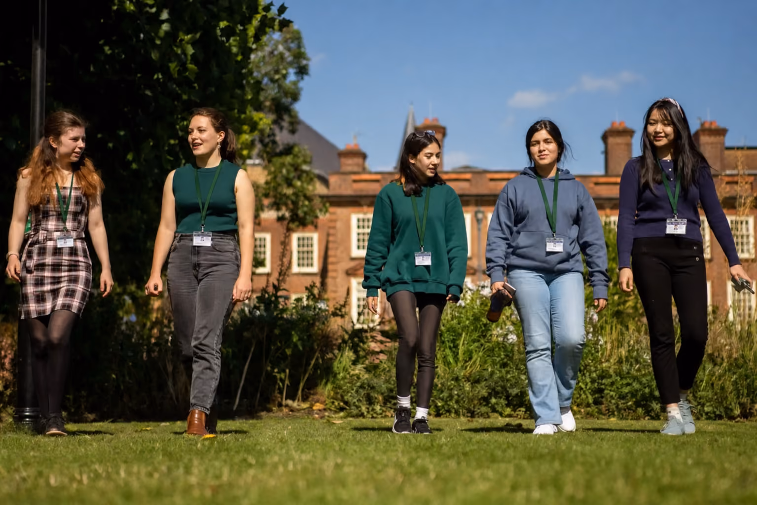 Five students walking across the lawn in front of a red-brick college building
