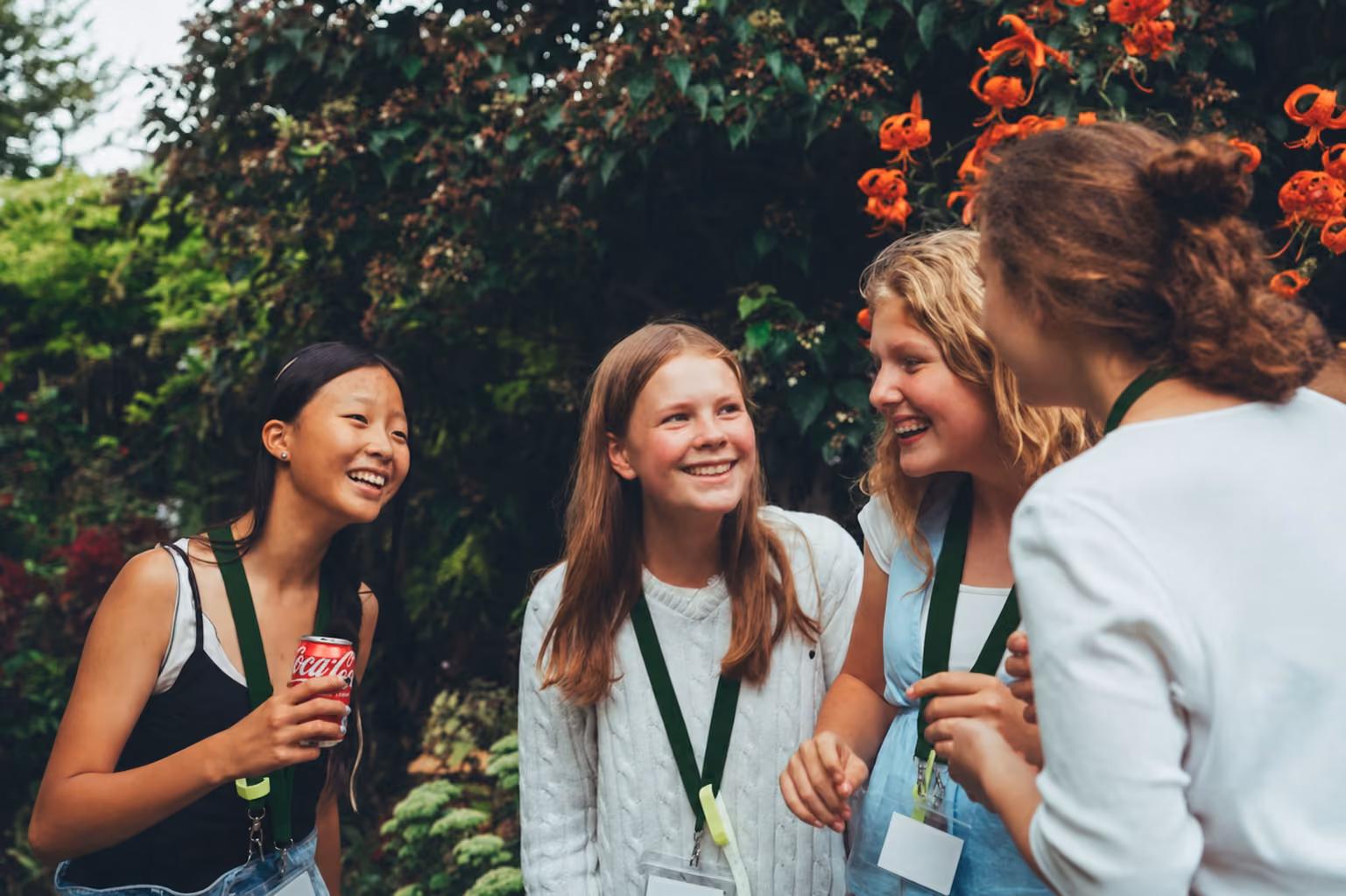 Four teenage girls smiling and chatting outdoors with greenery and orange flowers in the background.