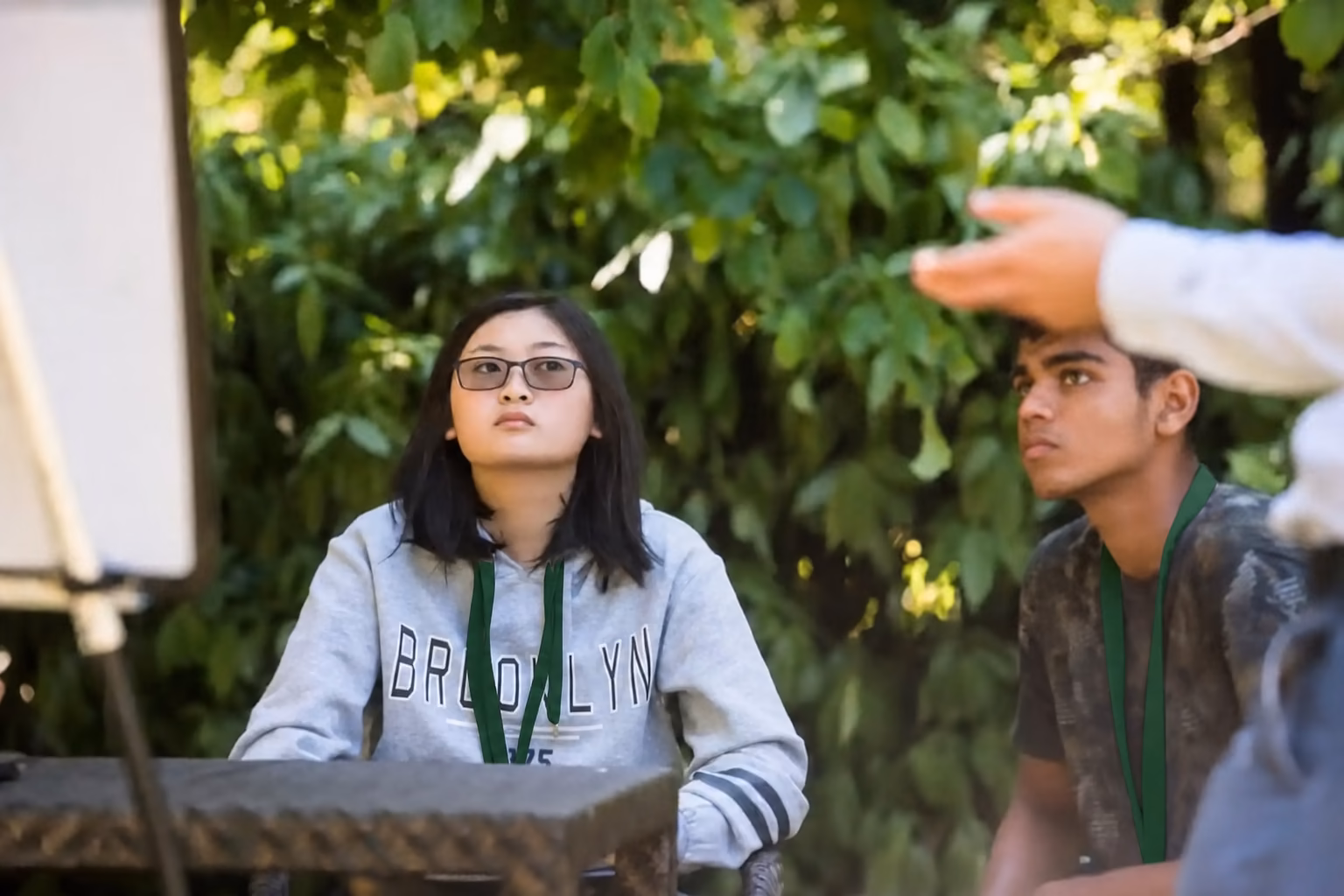 Two young people, a girl wearing glasses and a boy, attentively looking at a person gesturing outside the frame, outdoors with green foliage background.
