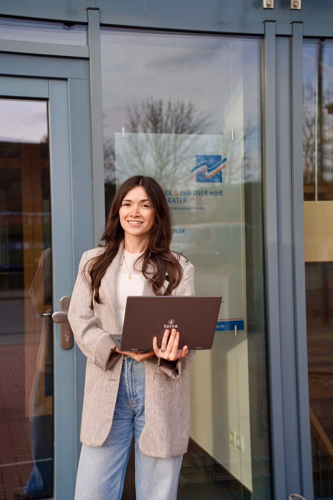 Lächelnde Frau steht mit Laptop vor einem Bürogebäude mit Glasfront.