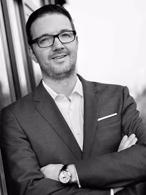 Schwarz-weiß-Porträt eines lächelnden Mannes mit Brille, der vor einem Gebäude im Anzug posiert.