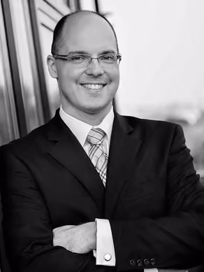 Ein lächelnder Mann in Anzug und Brille mit verschränkten Armen vor Fenstern.
