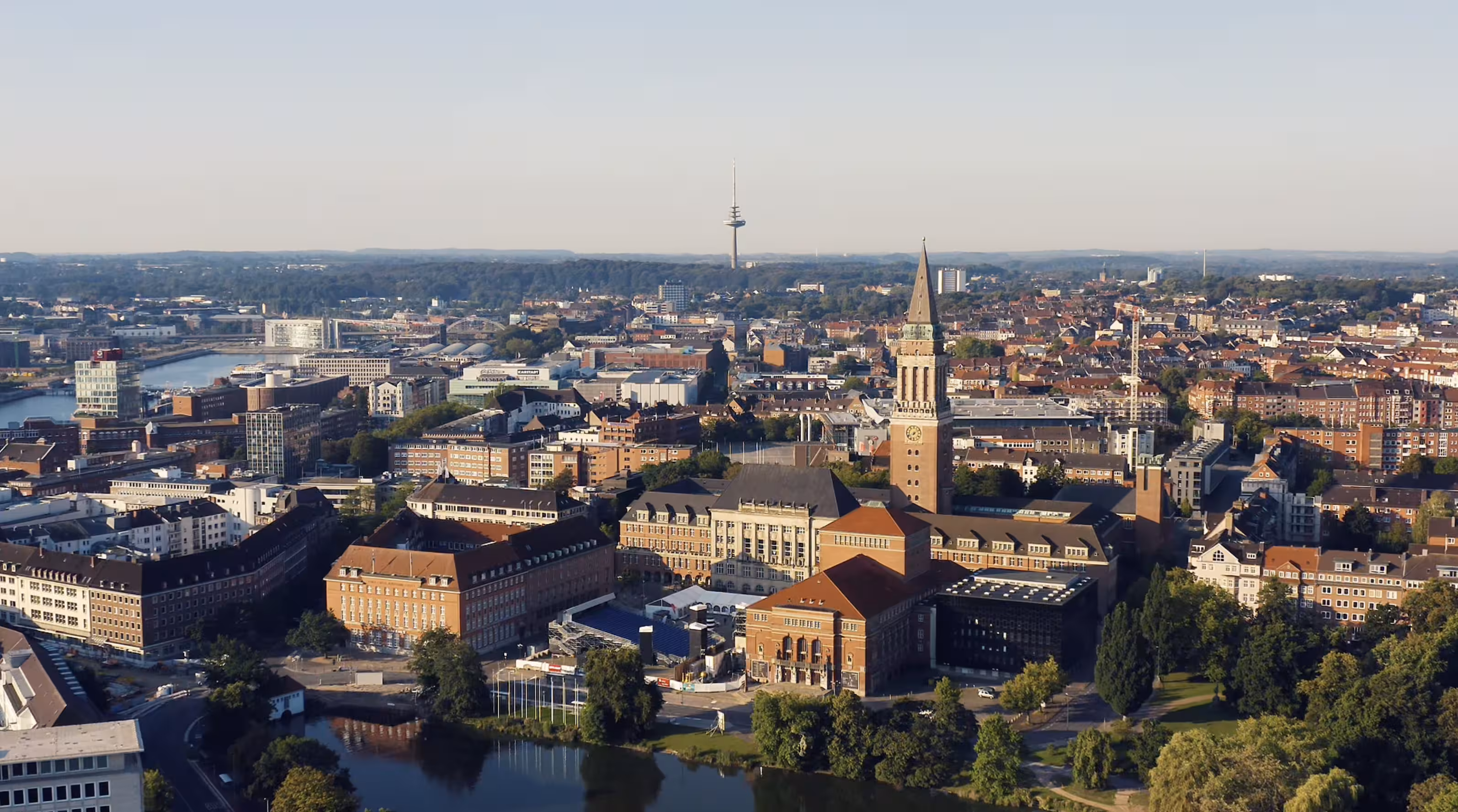 Luftaufnahme von Kiel mit Rathaus und Fluss, Gebäuden und Fernsehturm im Hintergrund.