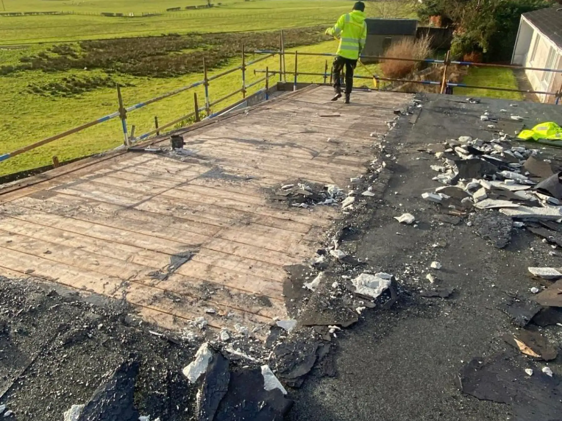 Worker in hi-vis on the Strathaven flat roof during strip-out, with scaffold edge protection and broken debris
