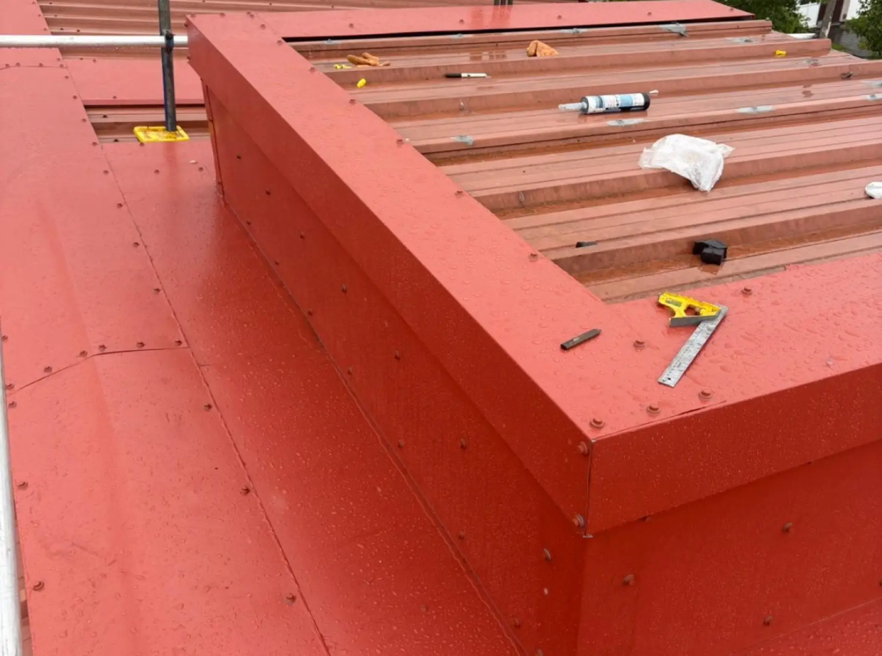 New red metal capping flashings around a roof upstand for Team Services, Glasgow, with a square and sealant left on the roof